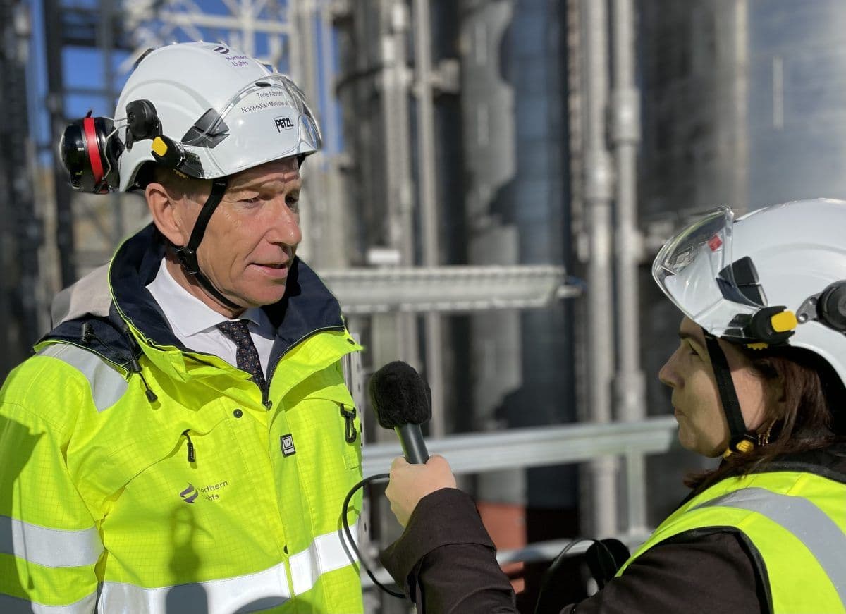 To personer i synlige jakker og vernehjelmer, den ene holder en mikrofon, ser ut til å gjennomføre et intervju på en industriplass.