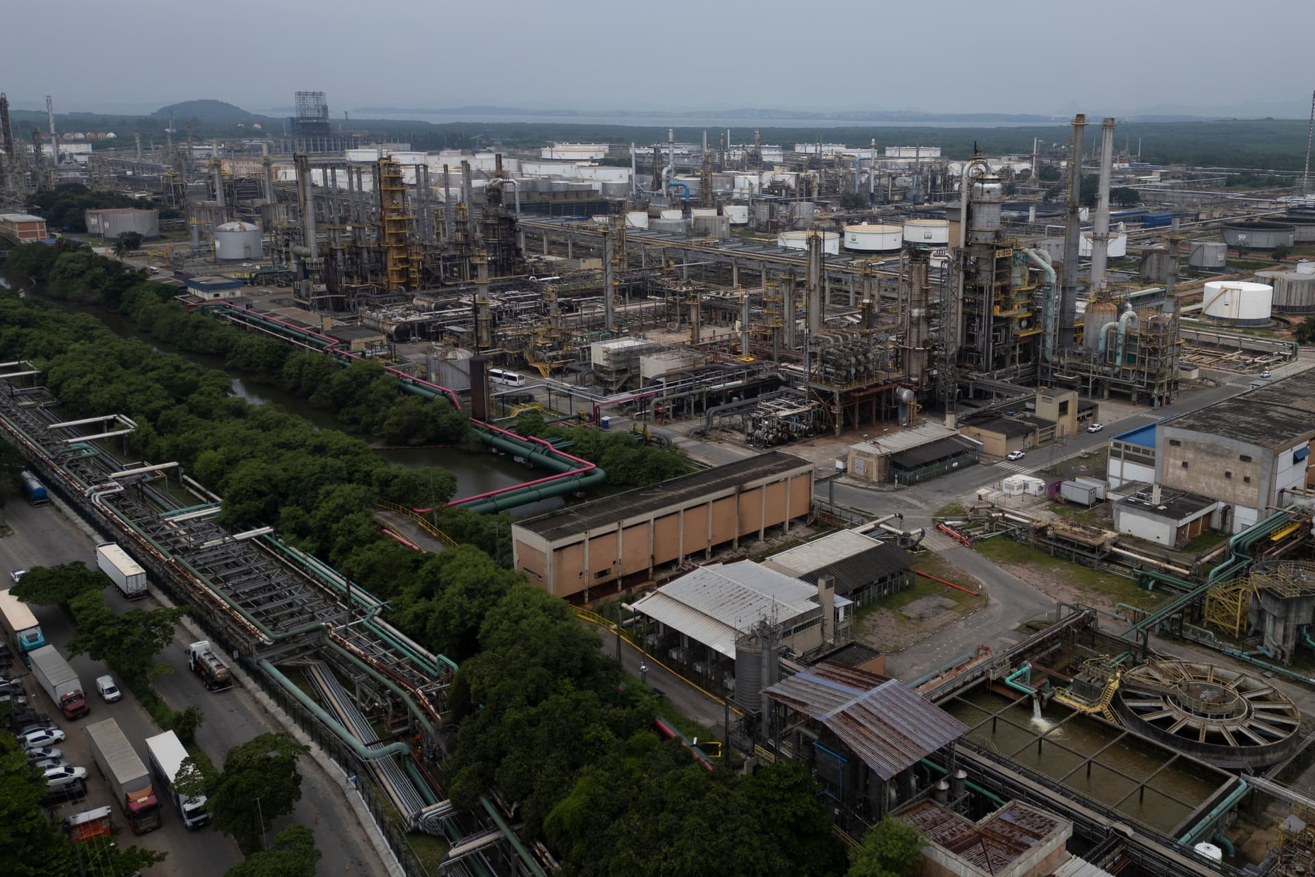 Luftfoto av et enormt industrianlegg med en rekke rør, tanker og konstruksjoner, ved siden av en trekantet kanal og en vei med lastebiler, under en overskyet himmel.