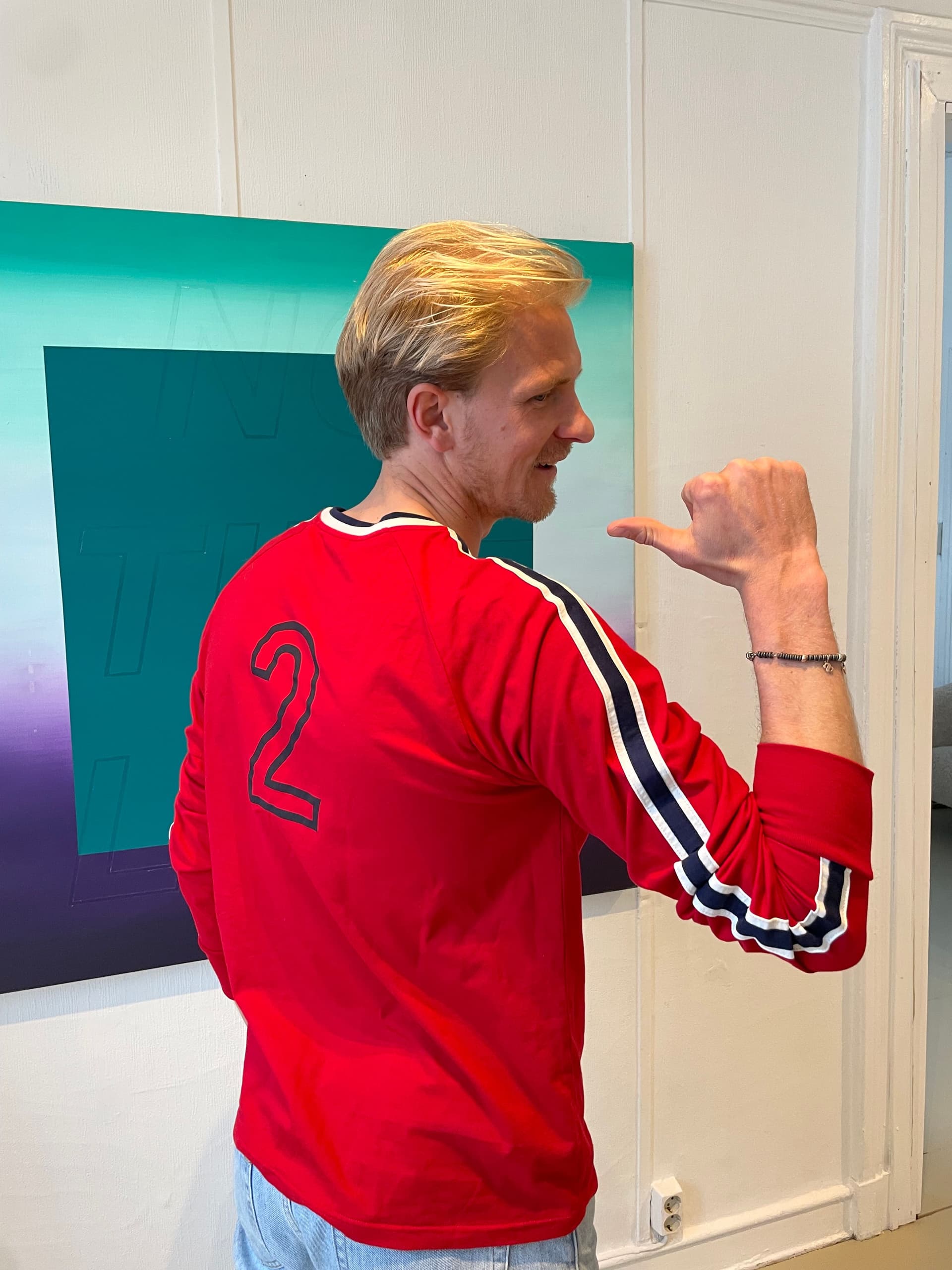 En blond mann i en rød trøye med tallet «2» på ryggen og stripete ermer smiler mens han gjør shaka-tegnet med høyre hånd.