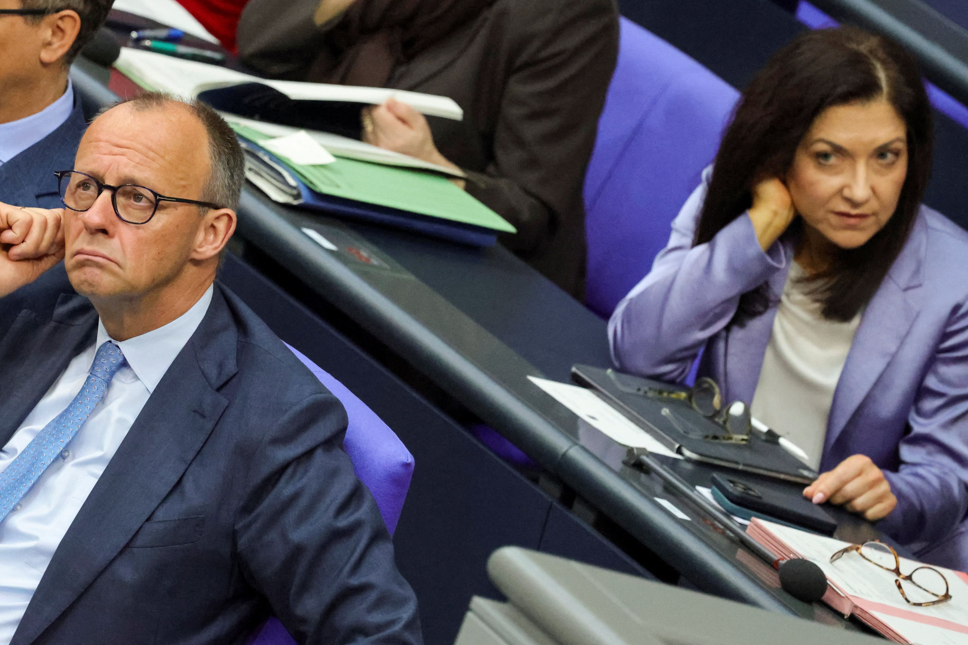 To politikere, en mann i dress som ser tankefullt opp, og en kvinne i en lilla blazer som ser rett frem med et alvorlig uttrykk, sittende ved pulter i en forsamlingssal.