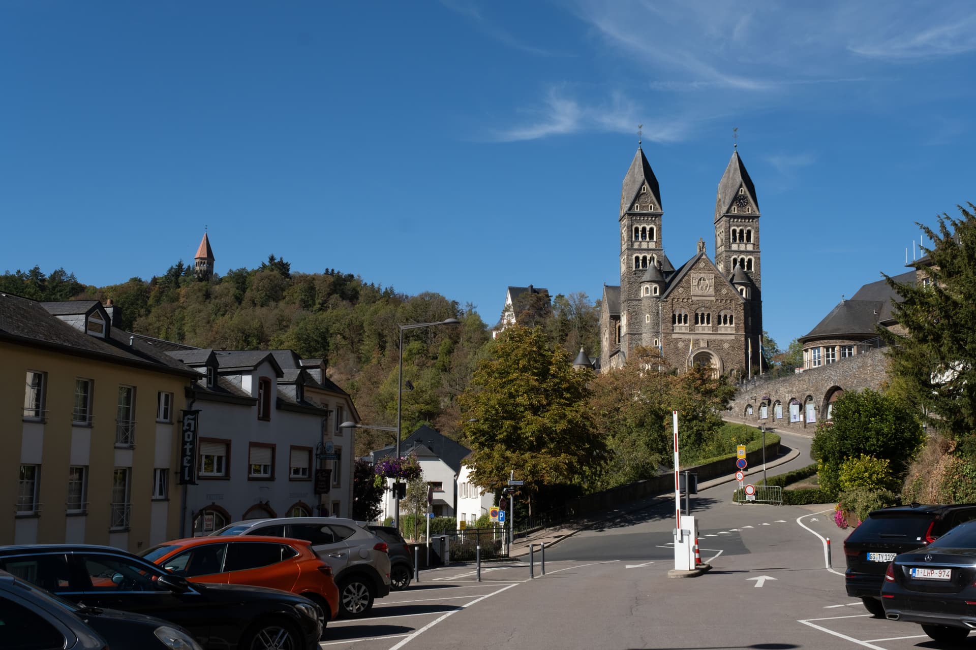 En storslått steinkirke med to spir ruver over en svingete gate med parkerte biler og bygninger, med en skogkledd ås i bakgrunnen under en blå himmel.