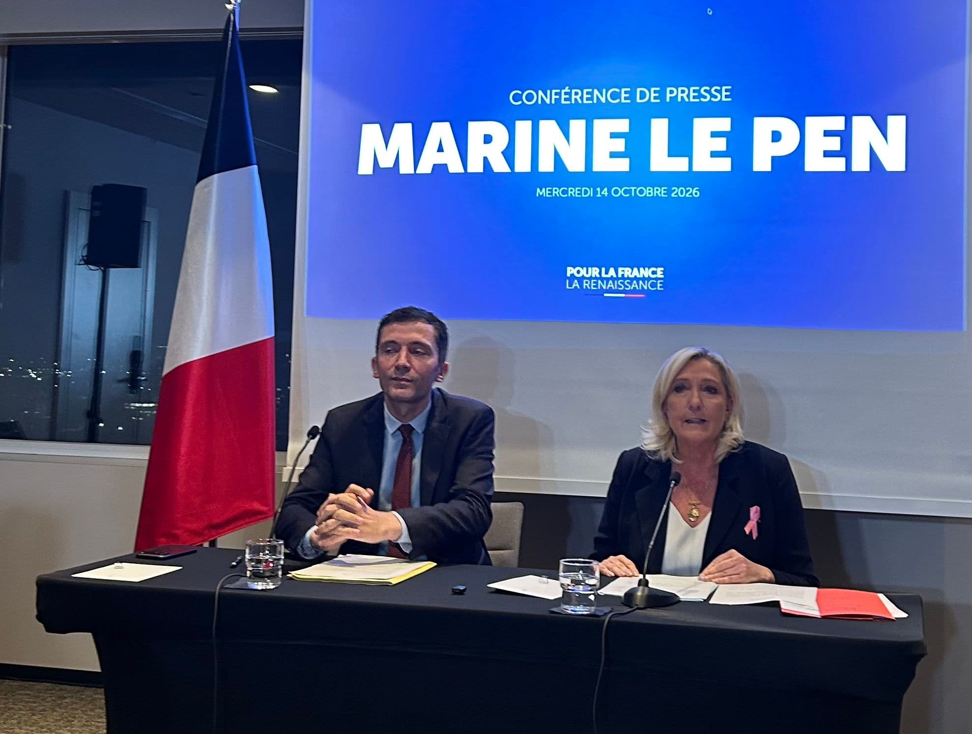 Marine Le Pen og en mann på en pressekonferanse med et fransk flagg og en skjerm som viser «Marine Le Pens pressekonferanse».