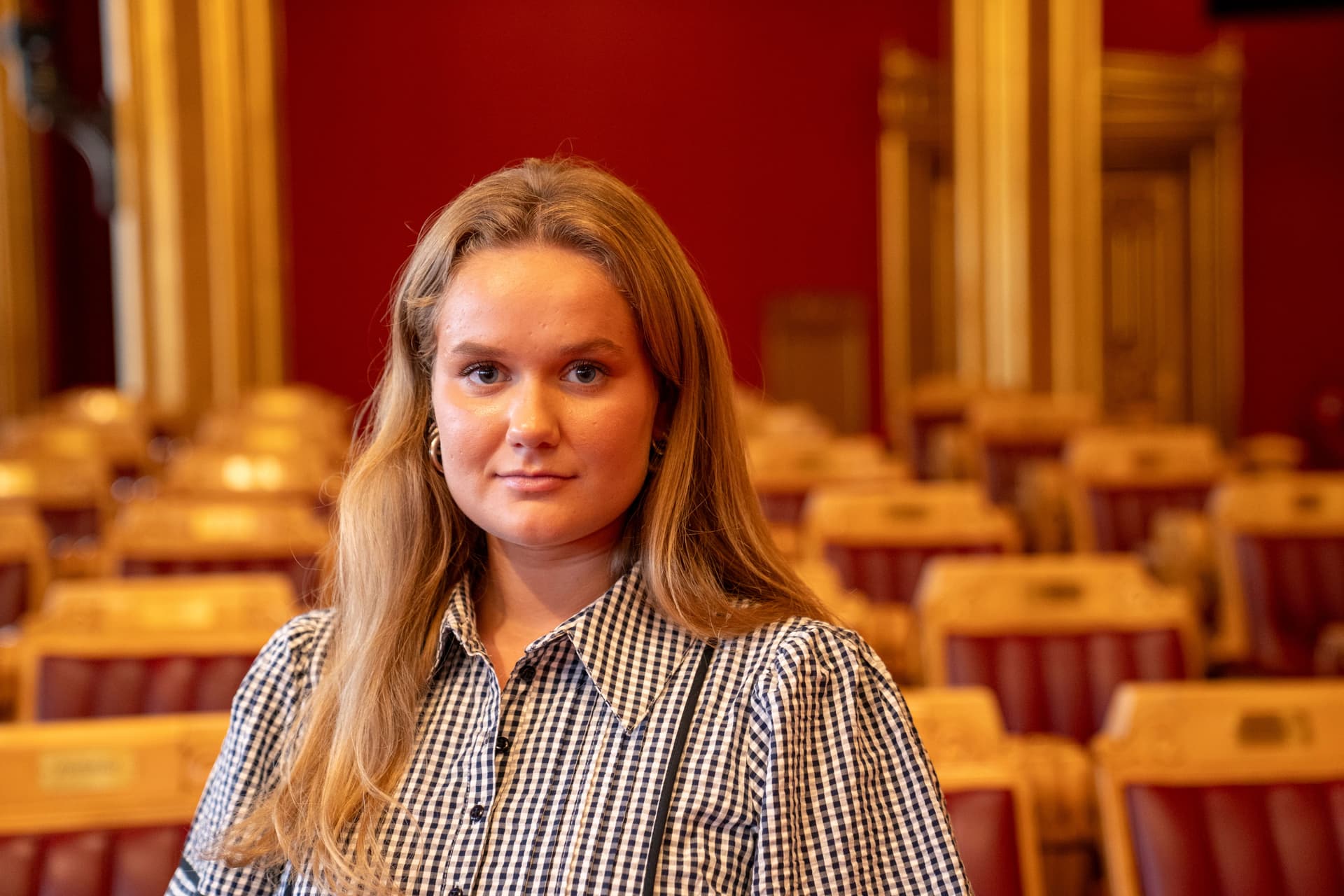 Ung kvinne i rutete skjorte i et utsmykket rom med sitteplasser i rødt og gull.