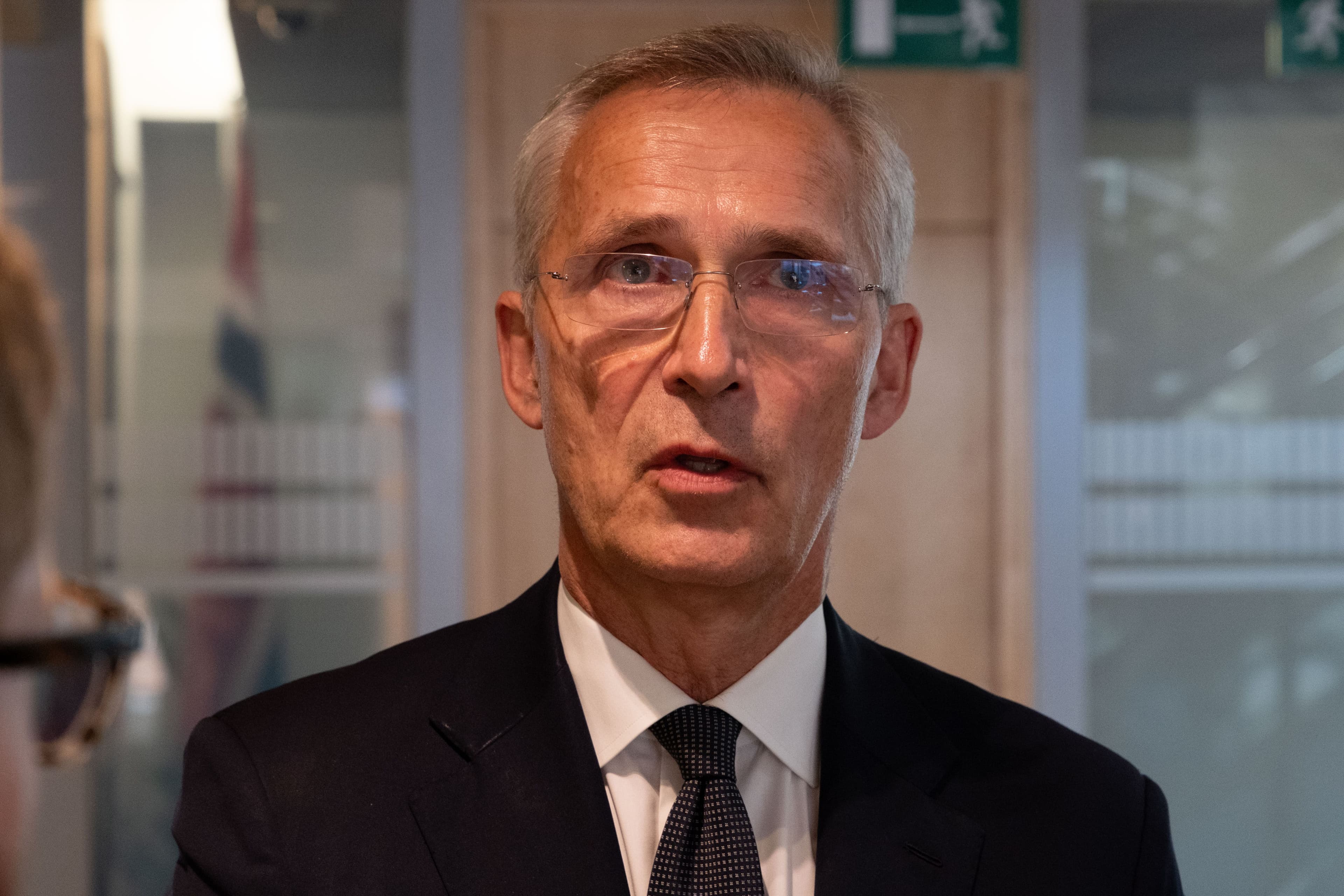 Jens Stoltenberg står foran en dør og et norsk flagg og ser opp mens han snakker.