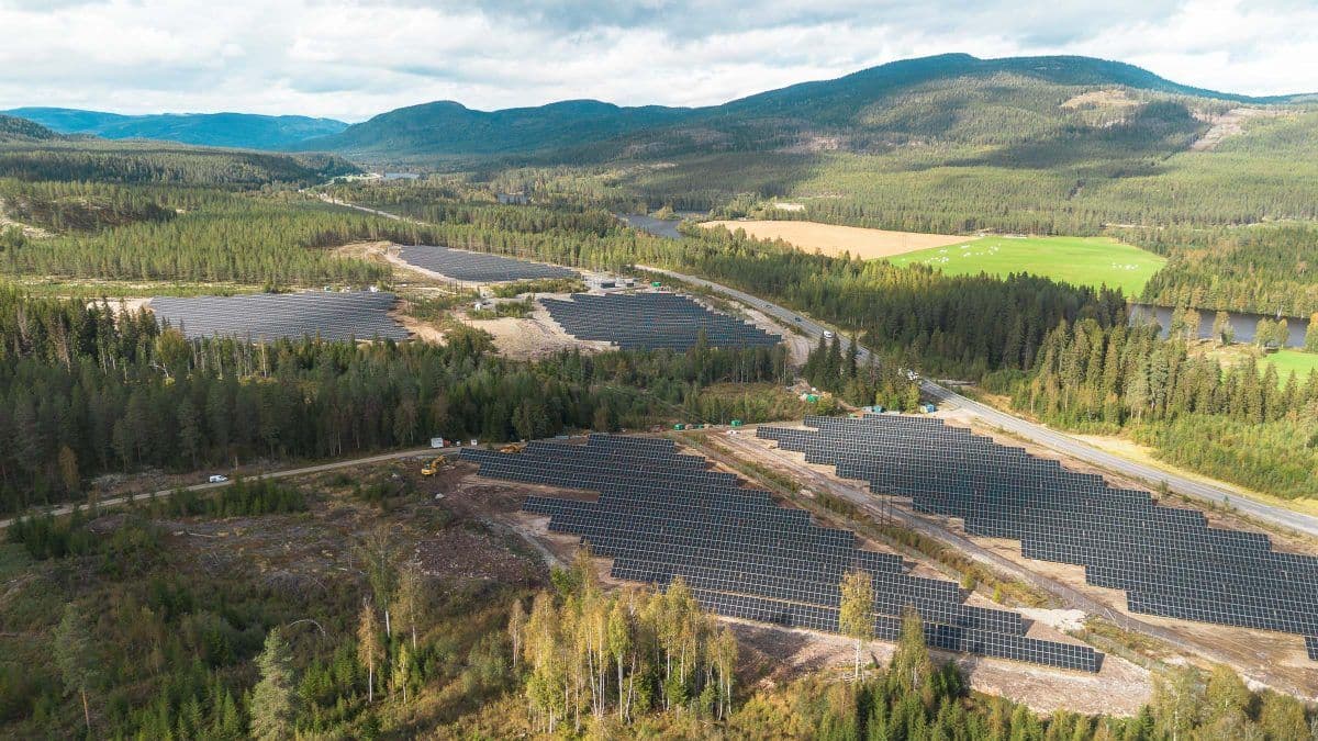 Luftfoto av flere solcellepanelinstallasjoner i et skogkledd, fjellrikt landskap under en delvis overskyet himmel.