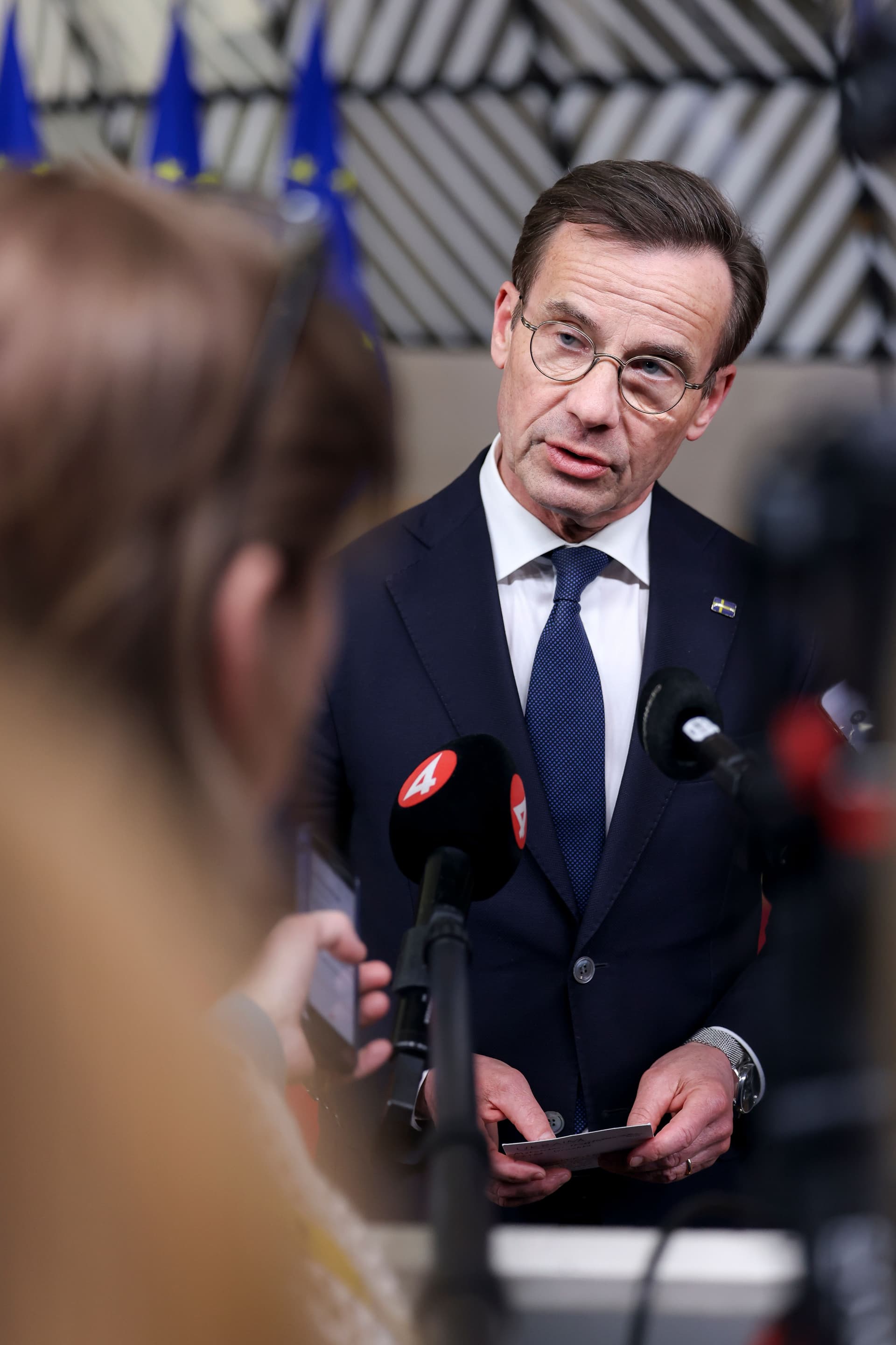 Mann i dress og briller som snakker i mikrofoner, med en svensk flaggnål og EU-flagg i bakgrunnen.