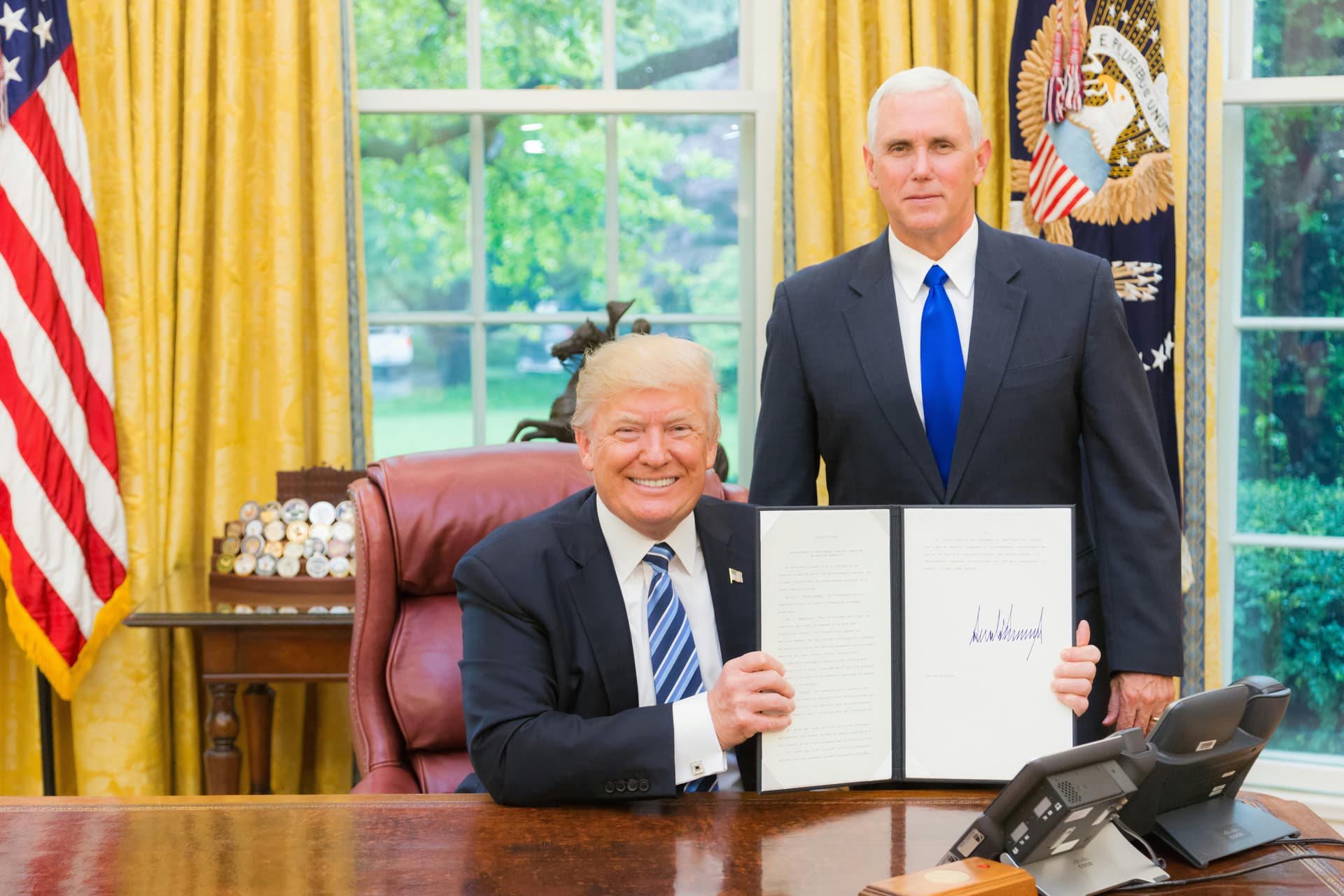 Donald Trump smiles while holding a signed document in the Oval Office, with Mike Pence standing behind him.