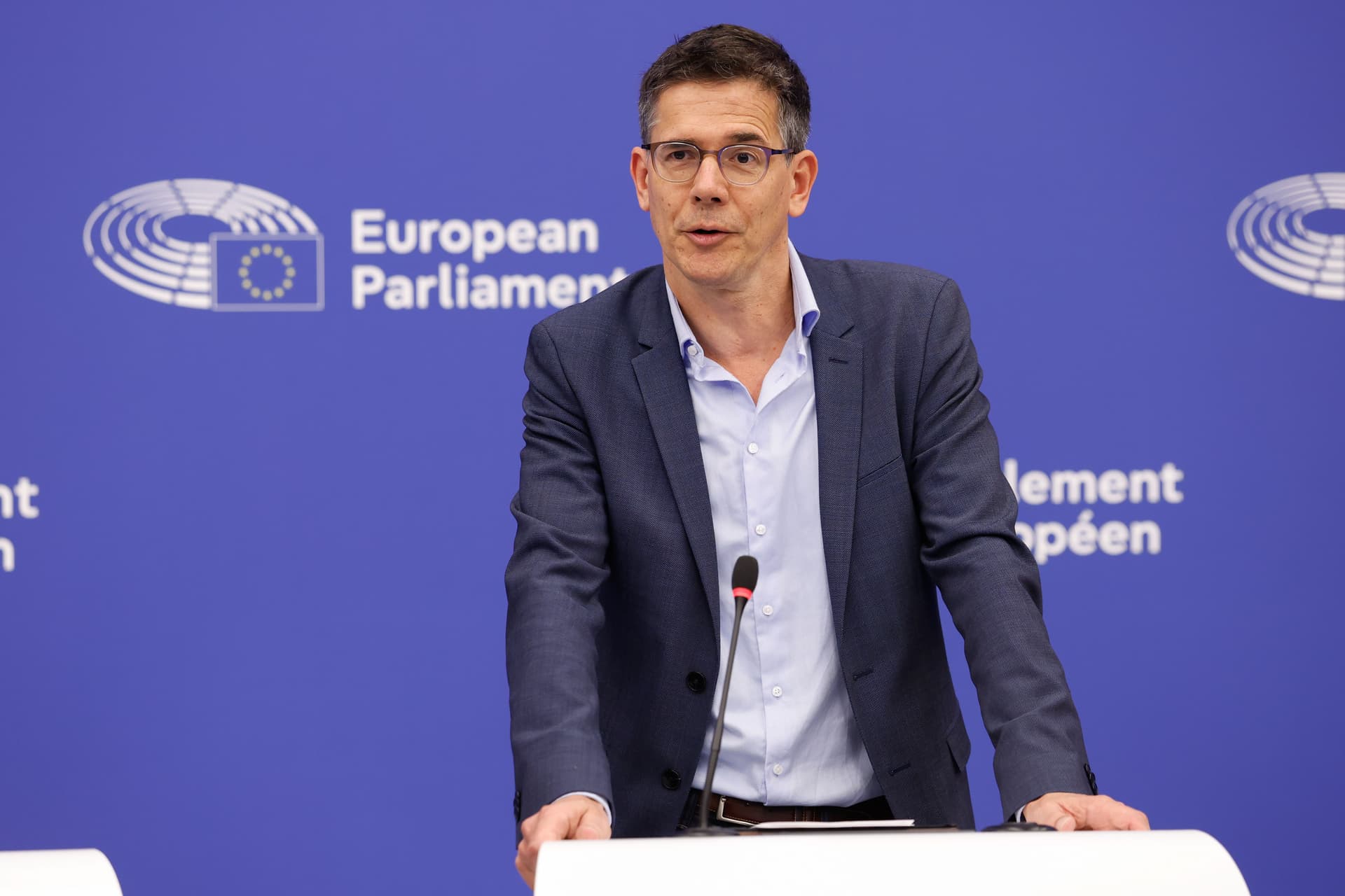 A man in a suit speaks at a podium with a European Parliament backdrop.