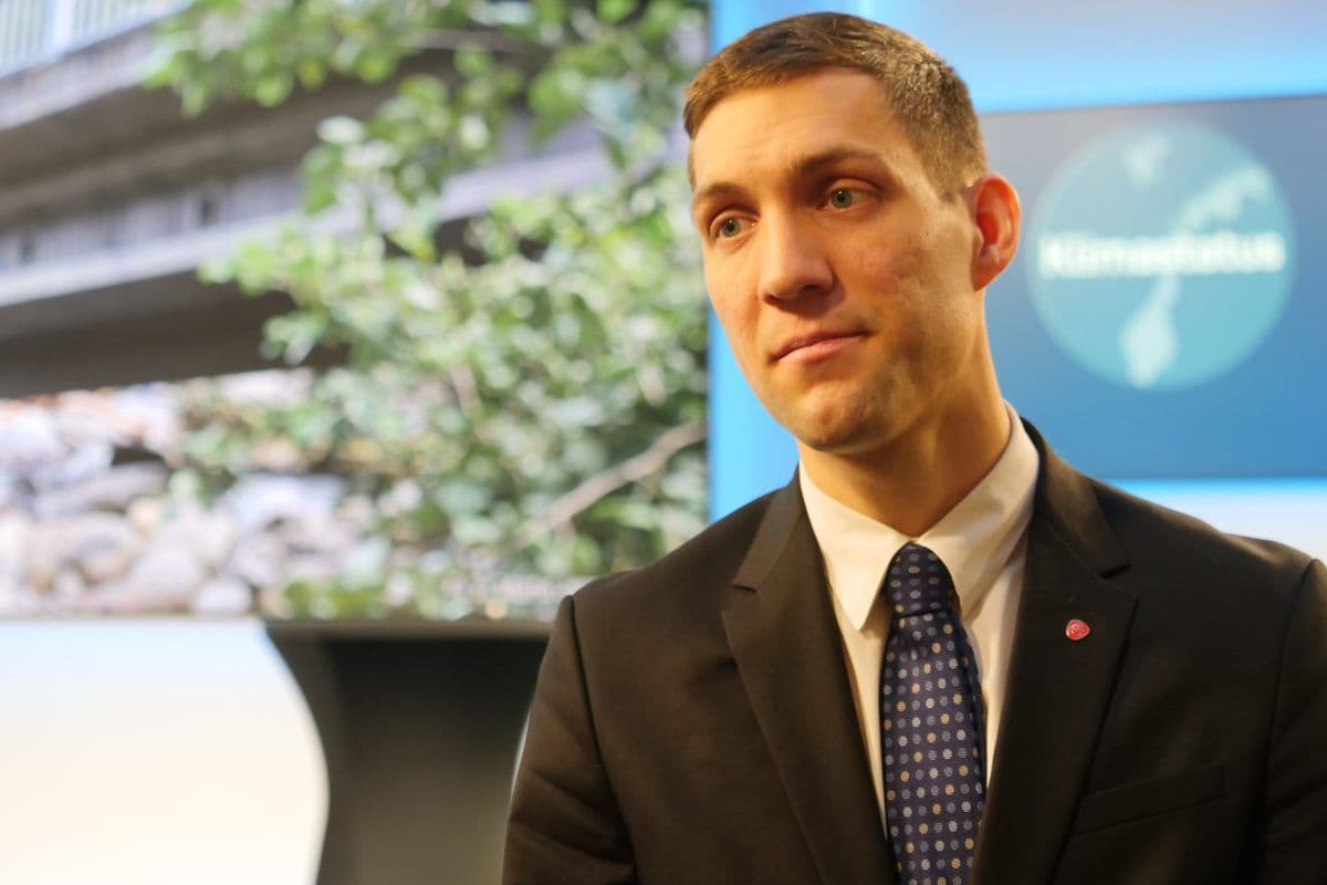 En mann i dress og slips står foran en skjerm som viser et naturbilde og en globusgrafikk merket "Klimastatus.