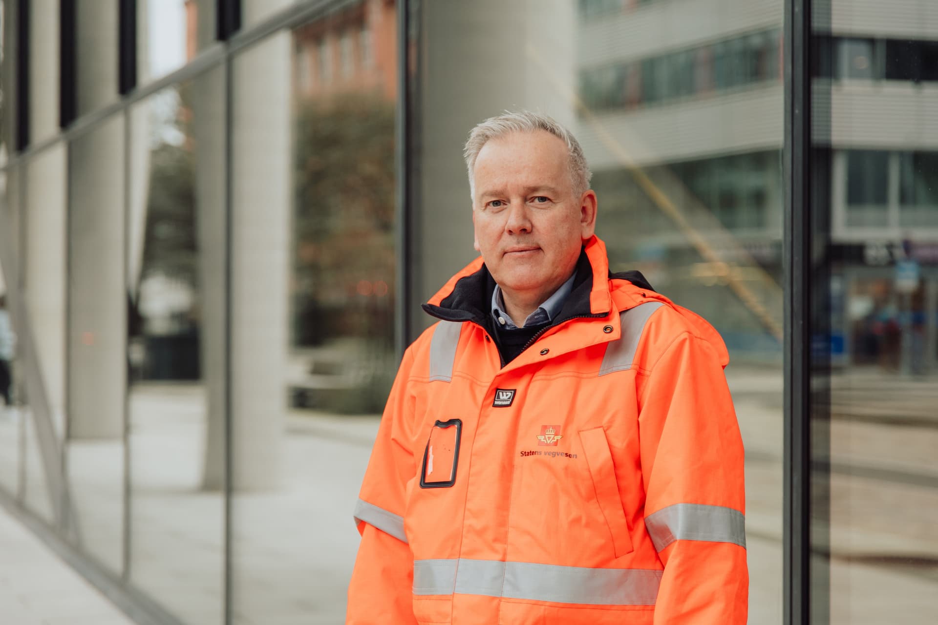 A man in an orange high-visibility jacket looks at the camera in front of a modern glass building.