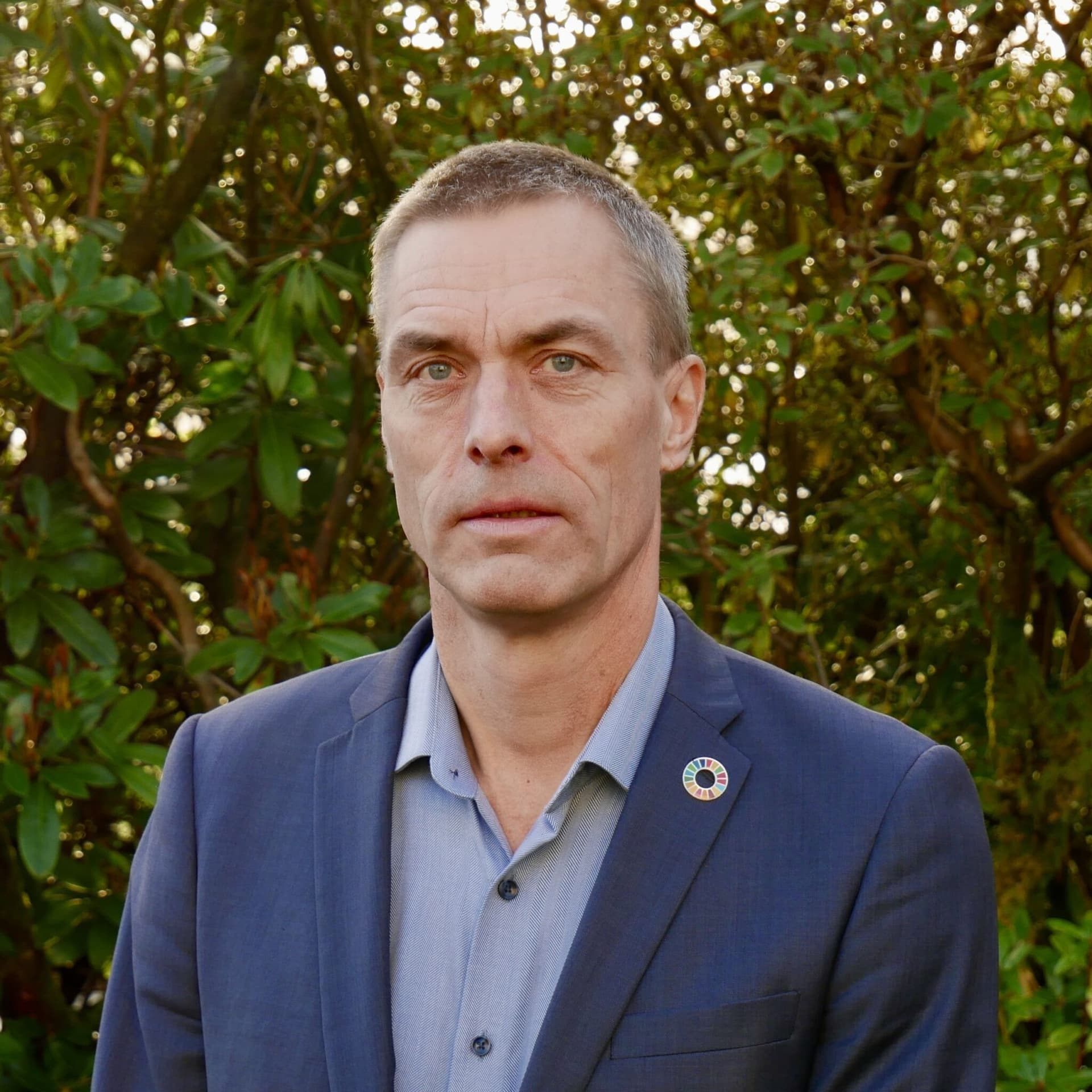 En mann med kort grått hår og blå øyne, iført en blå dress med en SDG-pin, står utendørs mot en bakgrunn av grønt løvverk.