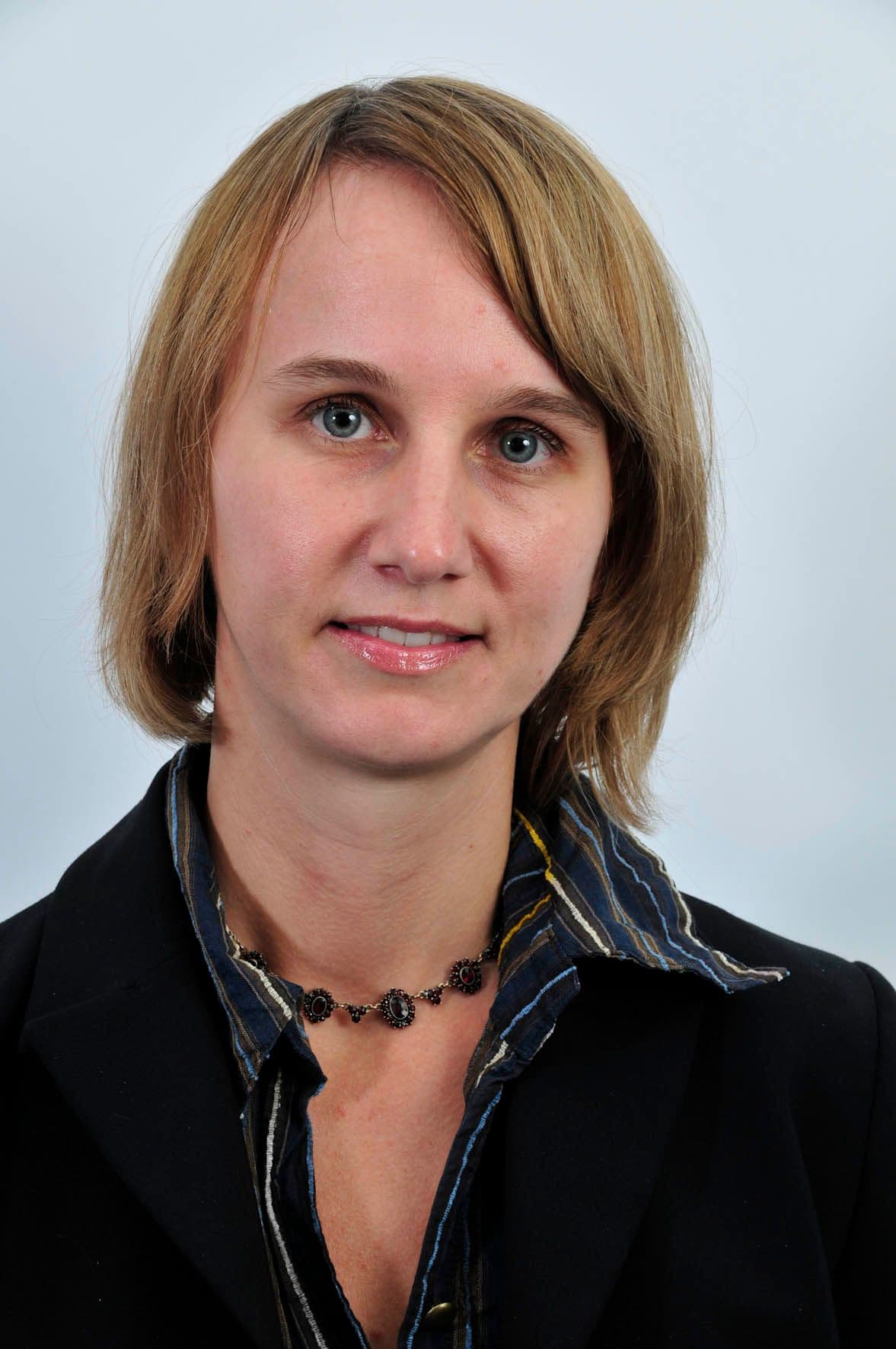 Portrett av en kvinne med kort blondt hår og blå øyne, iført en mørk blazer over en stripete skjorte og et halskjede, som ser rett inn i kameraet.