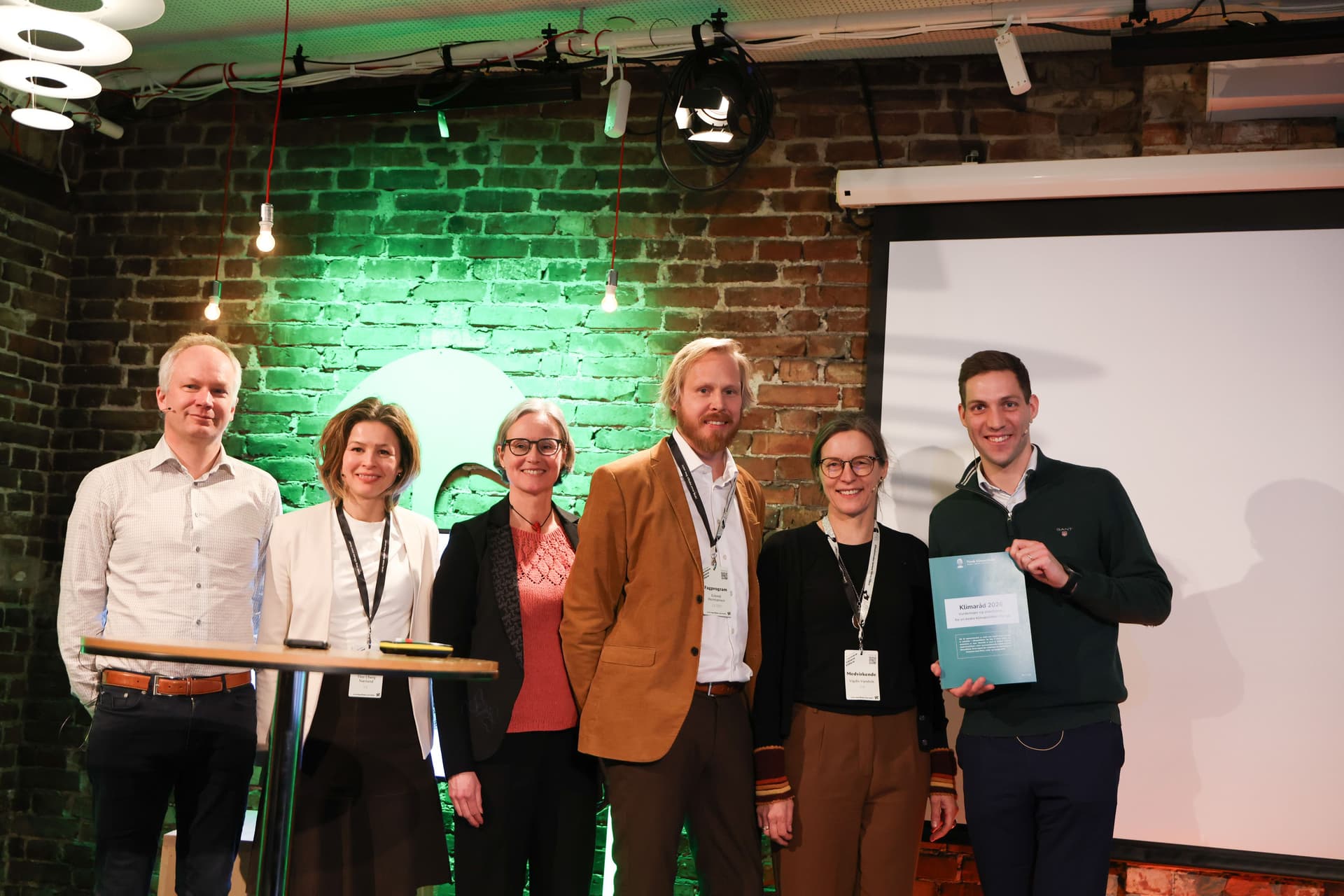 Tre menn og tre kvinner poserer på en scene, der en av mennene holder et sertifikat.