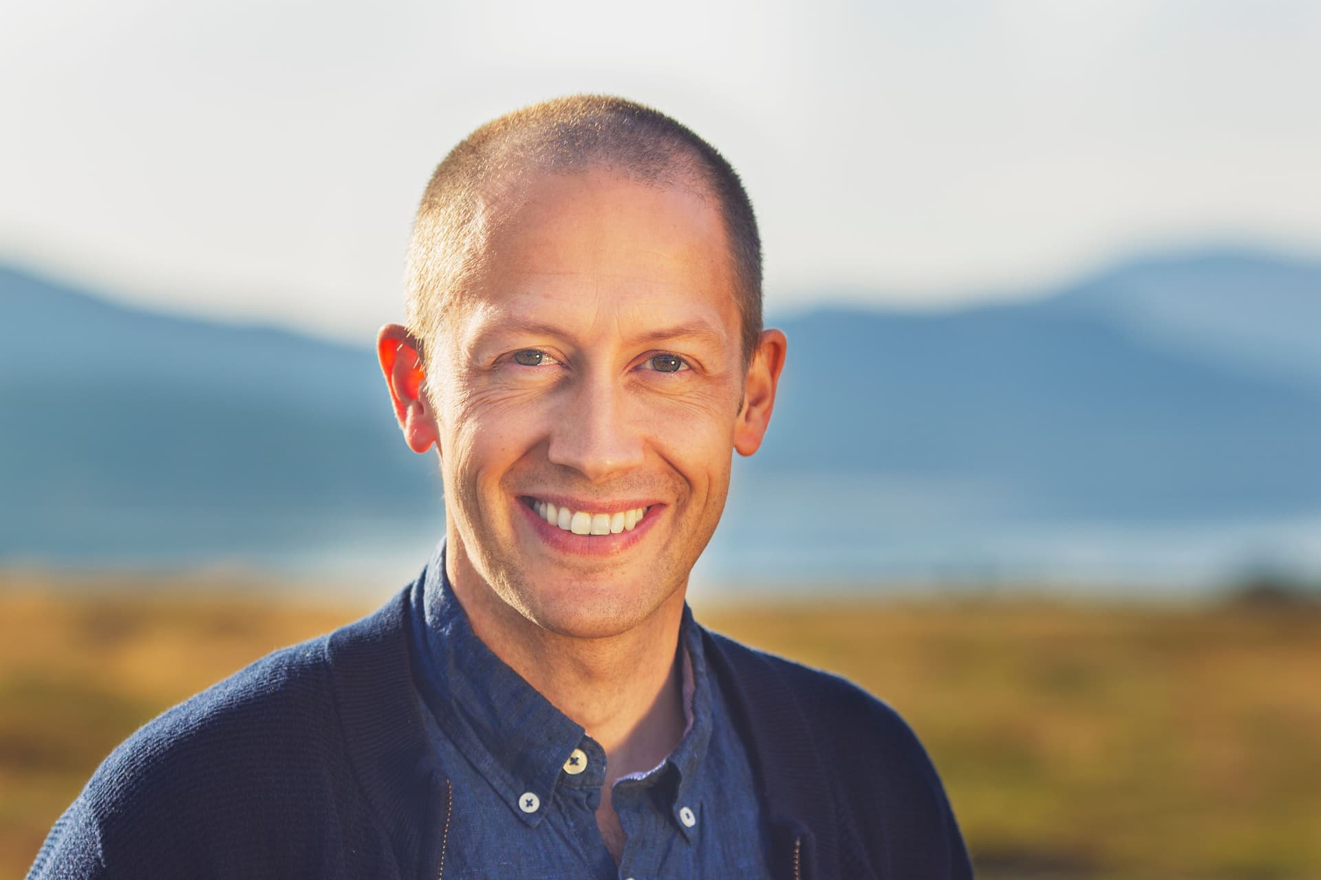 En smilende mann med barbert hode, iført en blå skjorte og mørk cardigan, står utendørs med fjell og gylne jorder i bakgrunnen.