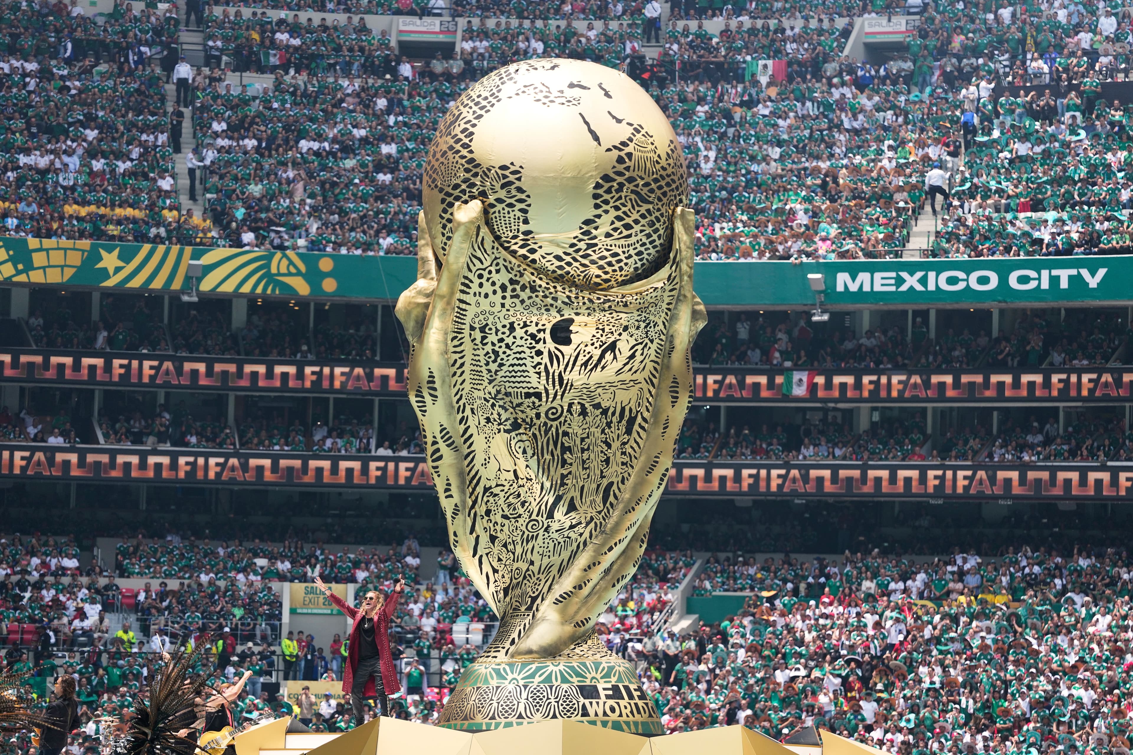 Et stort, utsmykket FIFA-VM-trofé i gull står på en scene i et fullsatt stadion med supportere kledd i grønt.