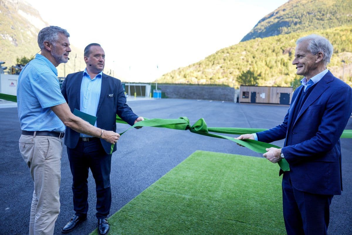 Tre menn står utendørs og forbereder seg på å klippe et stort grønt bånd som en del av en seremoni, med fjellene synlige i bakgrunnen.