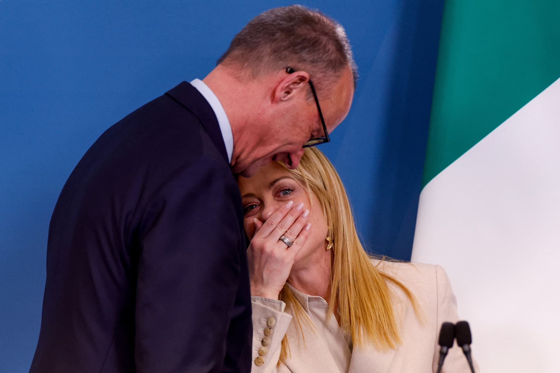 Giorgia Meloni covers her mouth with her hand while a man leans over and whispers to her.