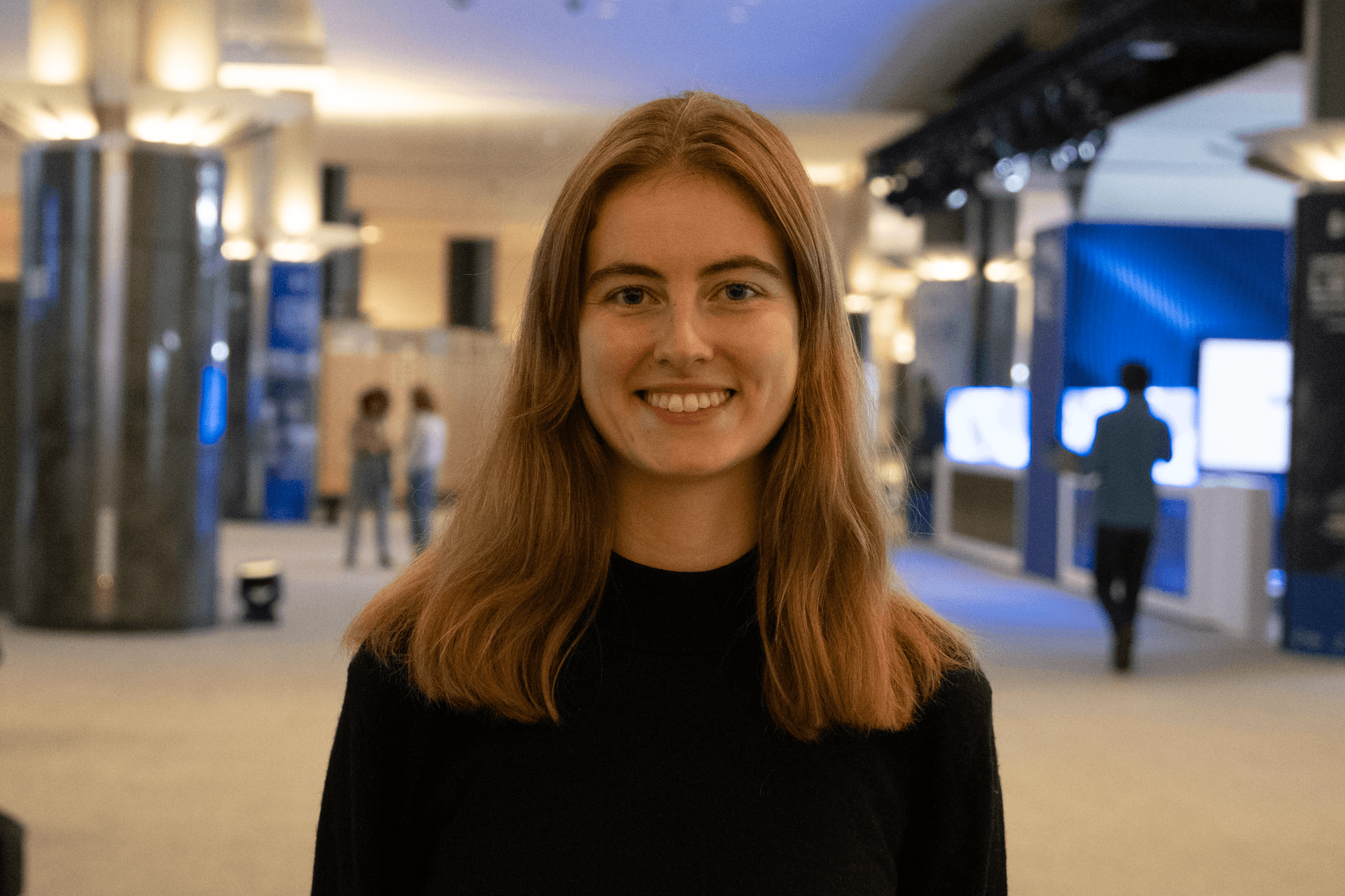 En smilende ung kvinne med langt, lysebrunt hår står i en moderne hall.