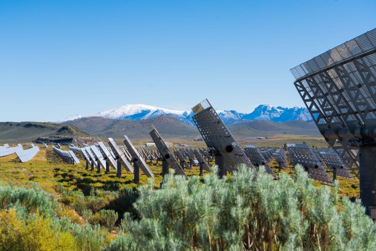 Rader av solcellepaneler er vinklet mot himmelen på en gresslette, med fjell i det fjerne og en klarblå himmel i bakgrunnen.