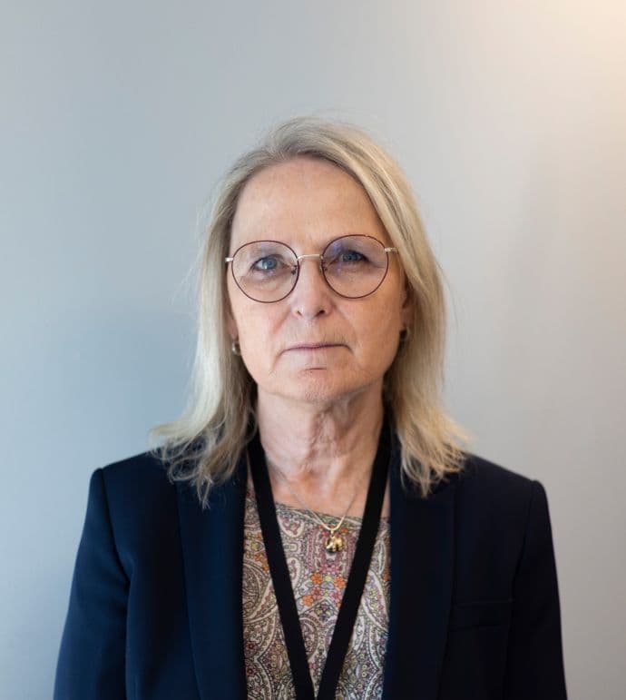 En kvinne med lyst hår og briller har på seg en mønstret topp og en mørk blazer, og ser inn i kameraet.