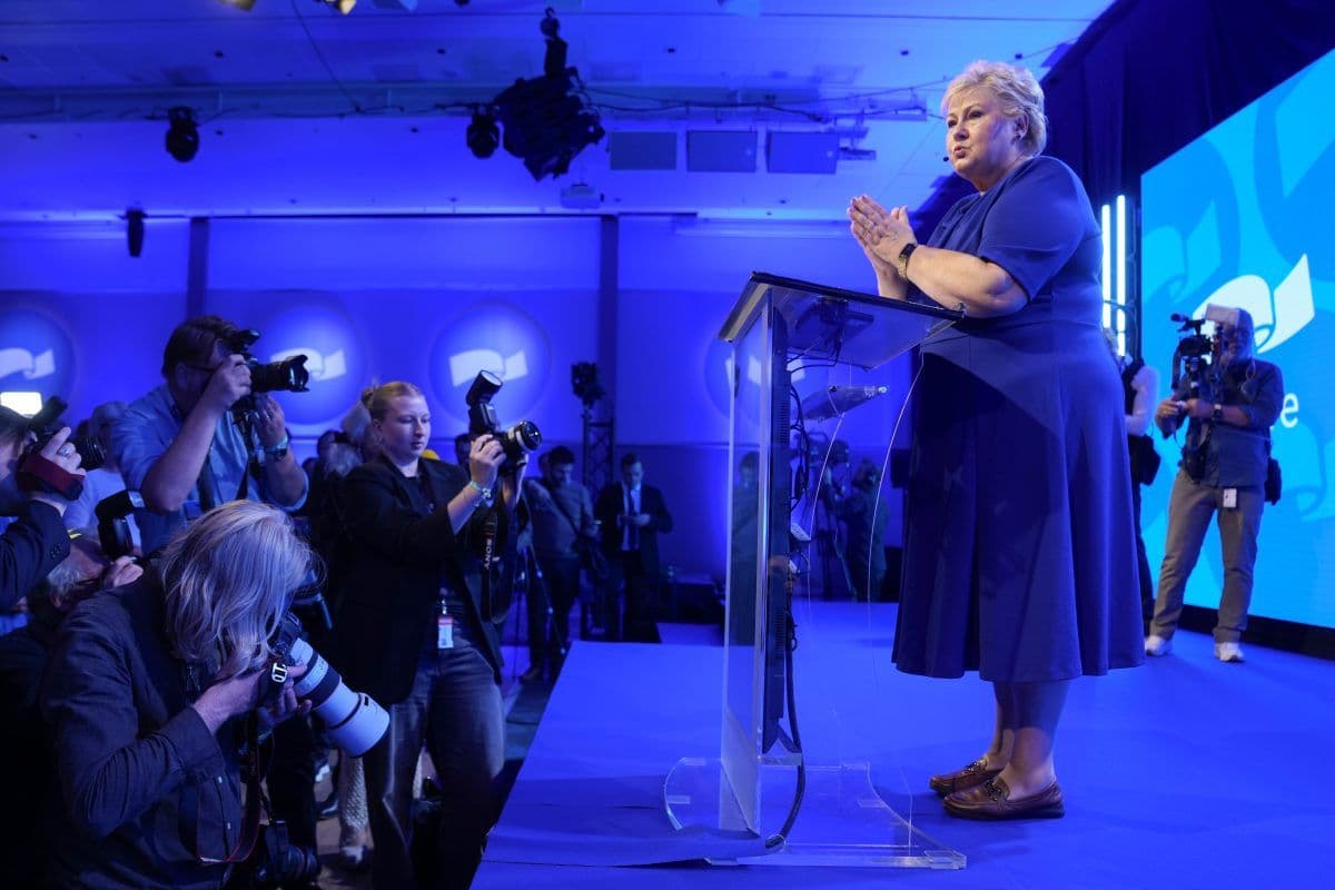 En kvinne i blå kjole taler på et tydelig podium på en scene, mens fotografer tar bilder i et rom med blå belysning.