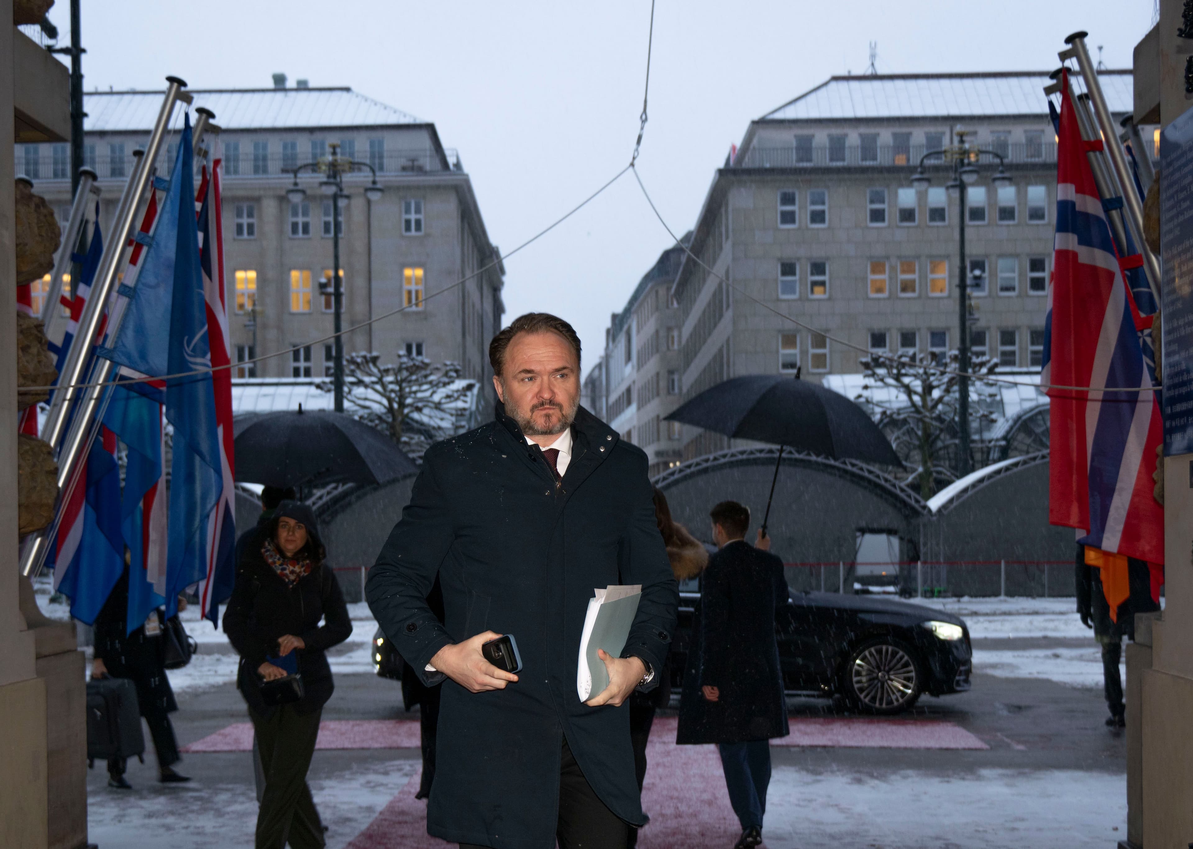 A man in a dark coat walks past NATO and Norwegian flags on a snowy street.