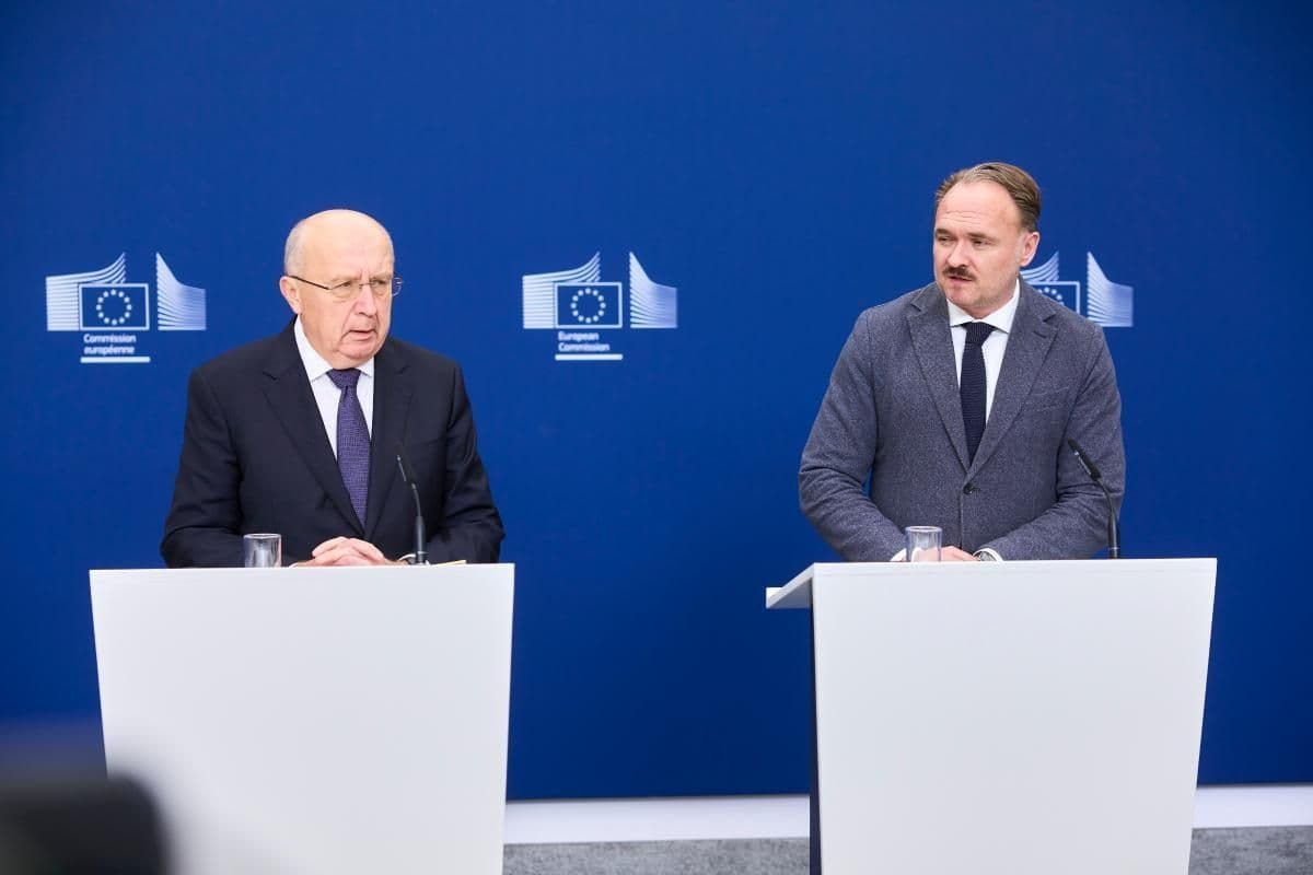 To menn i dress står ved talerstoler foran en blå bakgrunn med EU-kommisjonens logo.