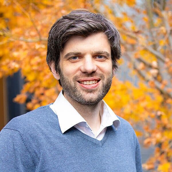 En mann med mørkt hår og skjegg, iført hvit skjorte og blå genser, står utendørs foran oransje høstløv.