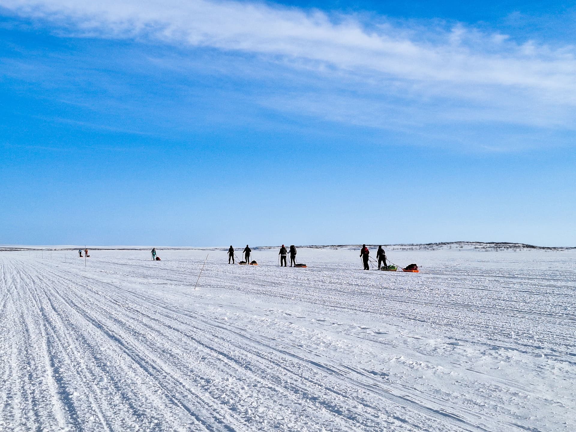 En gruppe mennesker som trekker sleder over en vidstrakt, snødekt slette med spor i snøen under en klar blå himmel.