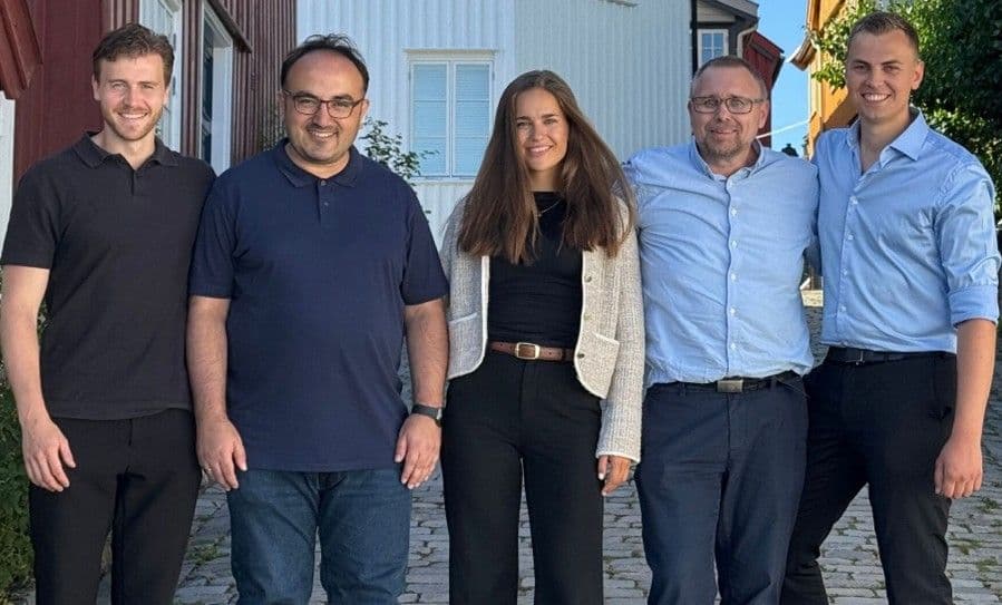 Five smiling people, four men and one woman, standing outdoors.