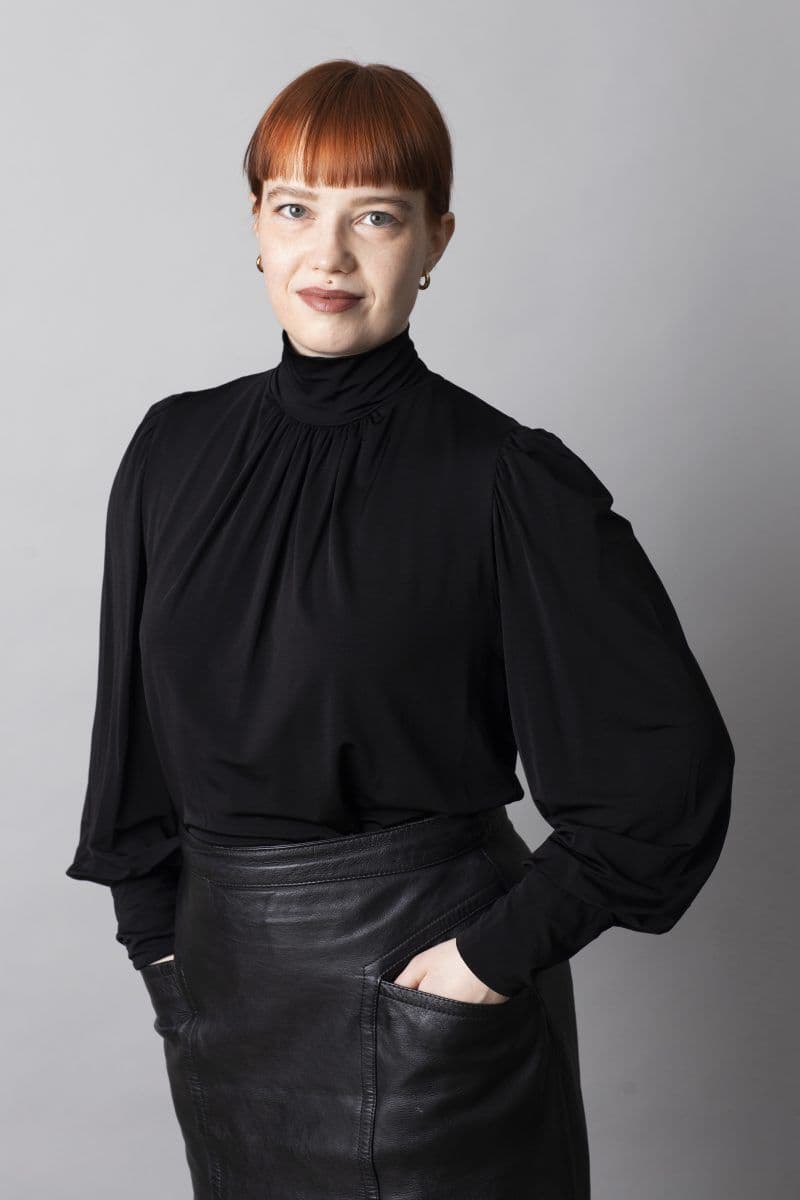 En person med rett, rødbrunt hår og lugg, iført svart langermet topp og svart skinnskjørt, står med hendene i lommene mot en ensfarget, lysegrå bakgrunn.