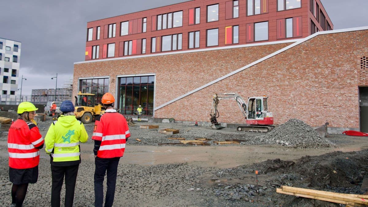 Fire bygningsarbeidere i sikkerhetsutstyr observerer en byggeplass med maskiner, grus og en moderne murbygning i bakgrunnen.