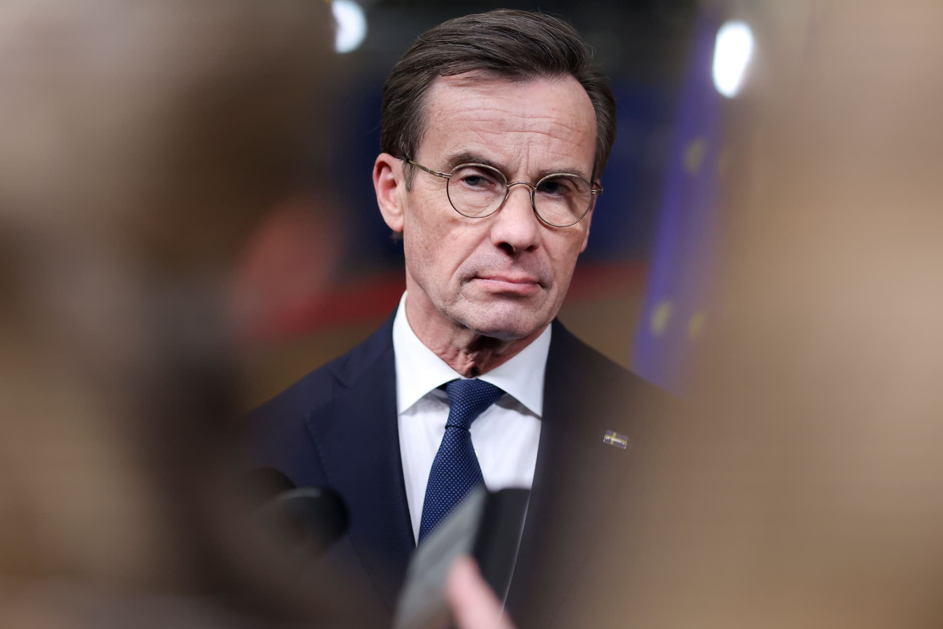 A man in a dark suit, blue tie, and glasses with a serious expression, wearing a Swedish flag pin on his lapel.