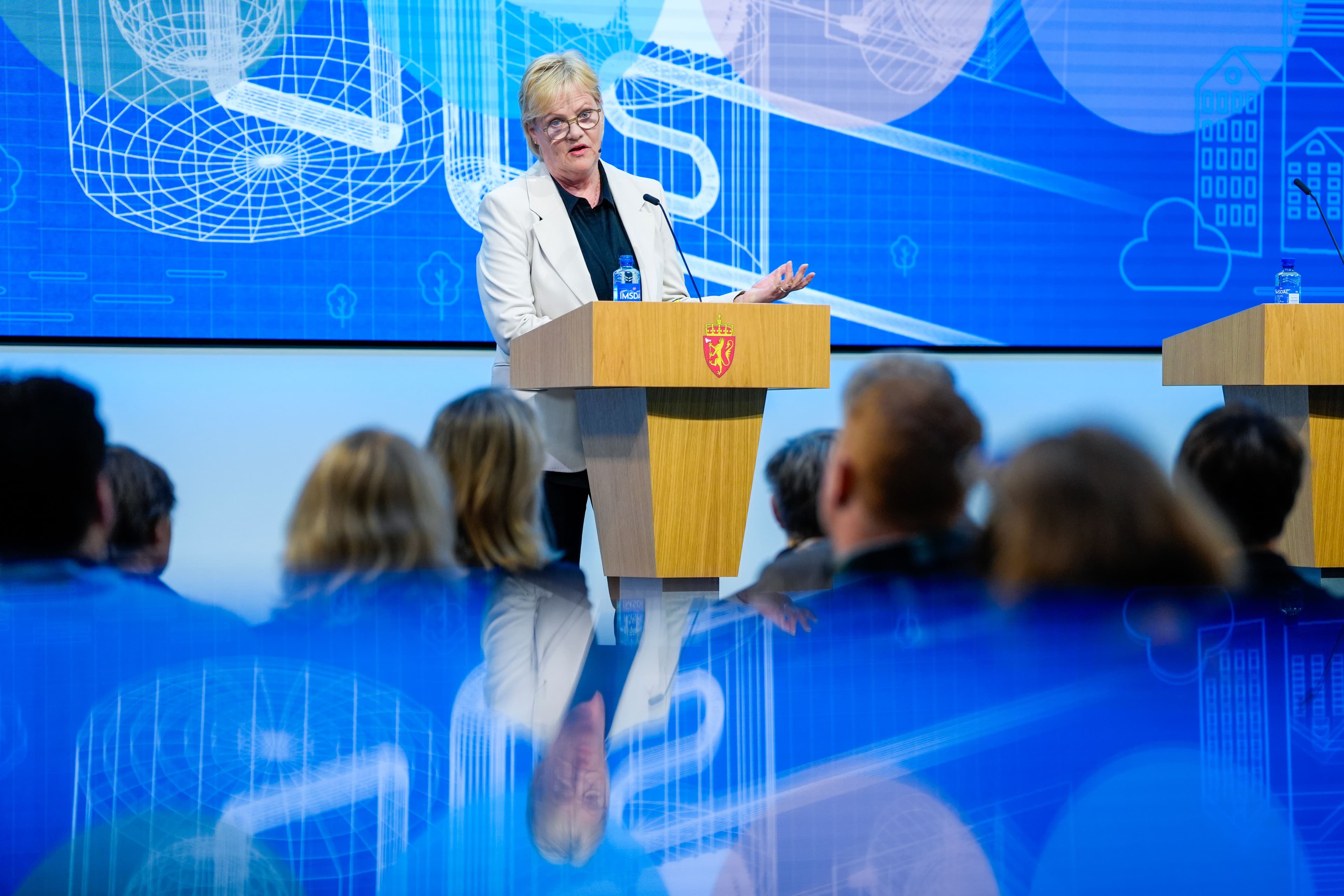 En kvinne i hvit blazer taler fra en talerstol med et statlig våpenskjold, mot en blå digital bakgrunn, med et publikum uskarpt i forgrunnen.