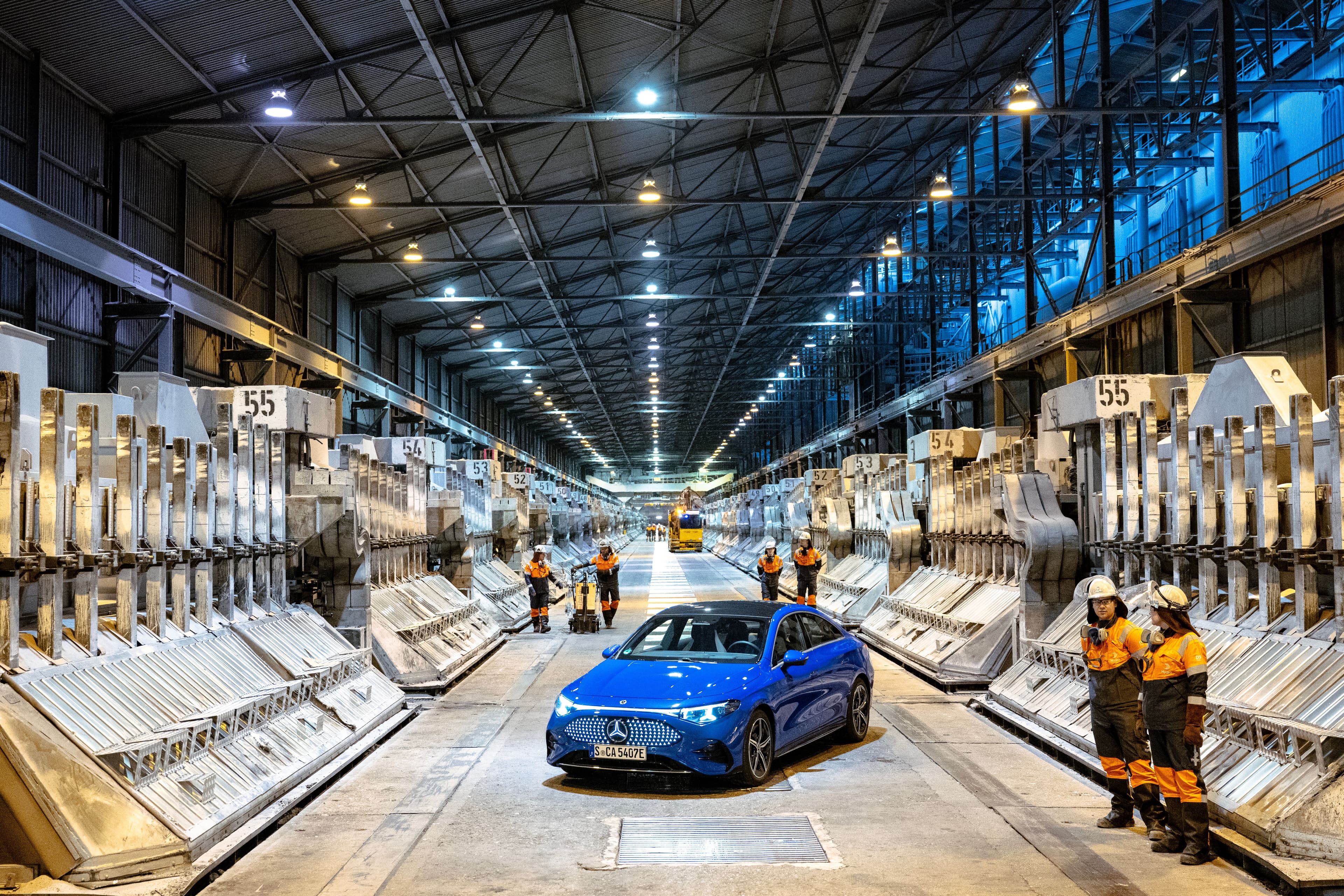 En blå Mercedes-Benz som står parkert i en stor industrisal med rekker av maskiner og flere arbeidere i verneutstyr.