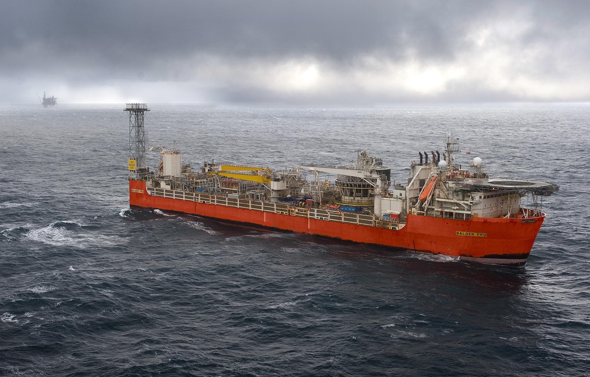 Et oransje offshore produksjonsskip på urolig sjø under en skyet himmel, med en oljerigg i det fjerne.
