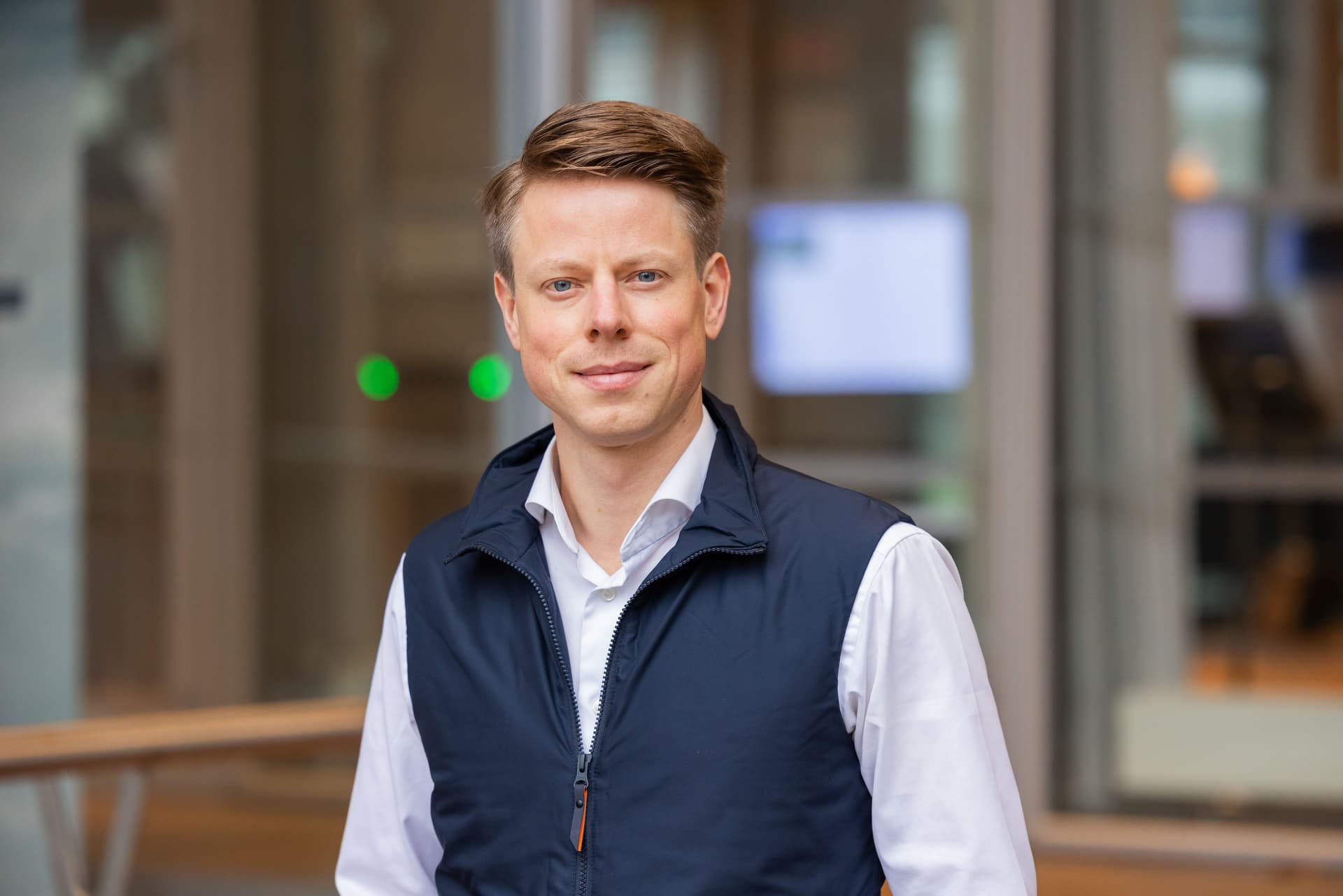 En mann med lysebrunt hår smiler, iført en hvit skjorte og en mørkeblå vest.