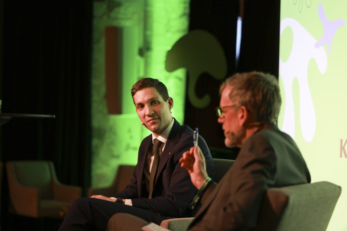 To menn i dress sitter og er i samtale på en scene med et grønt bakteppe som viser abstrakte former. En mann gestikulerer med en penn.