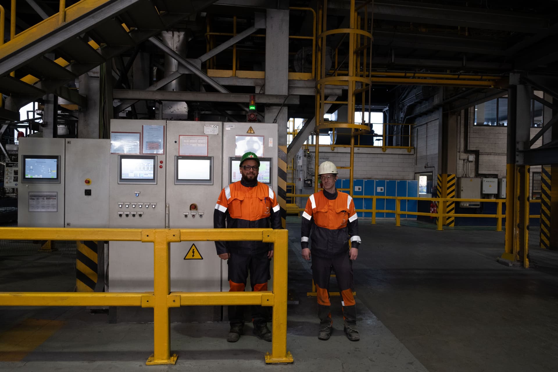 To arbeidere i oransje og svarte synlighetsklær, den ene med caps og den andre med vernehjelm, står i et industrianlegg ved et kontrollpanel og et gult sikkerhetsrekkverk.