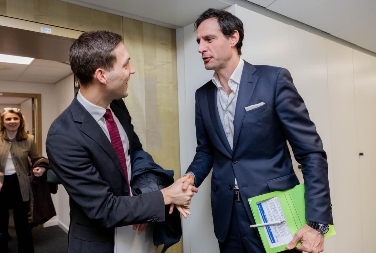 Two men in suits shake hands in a hallway.