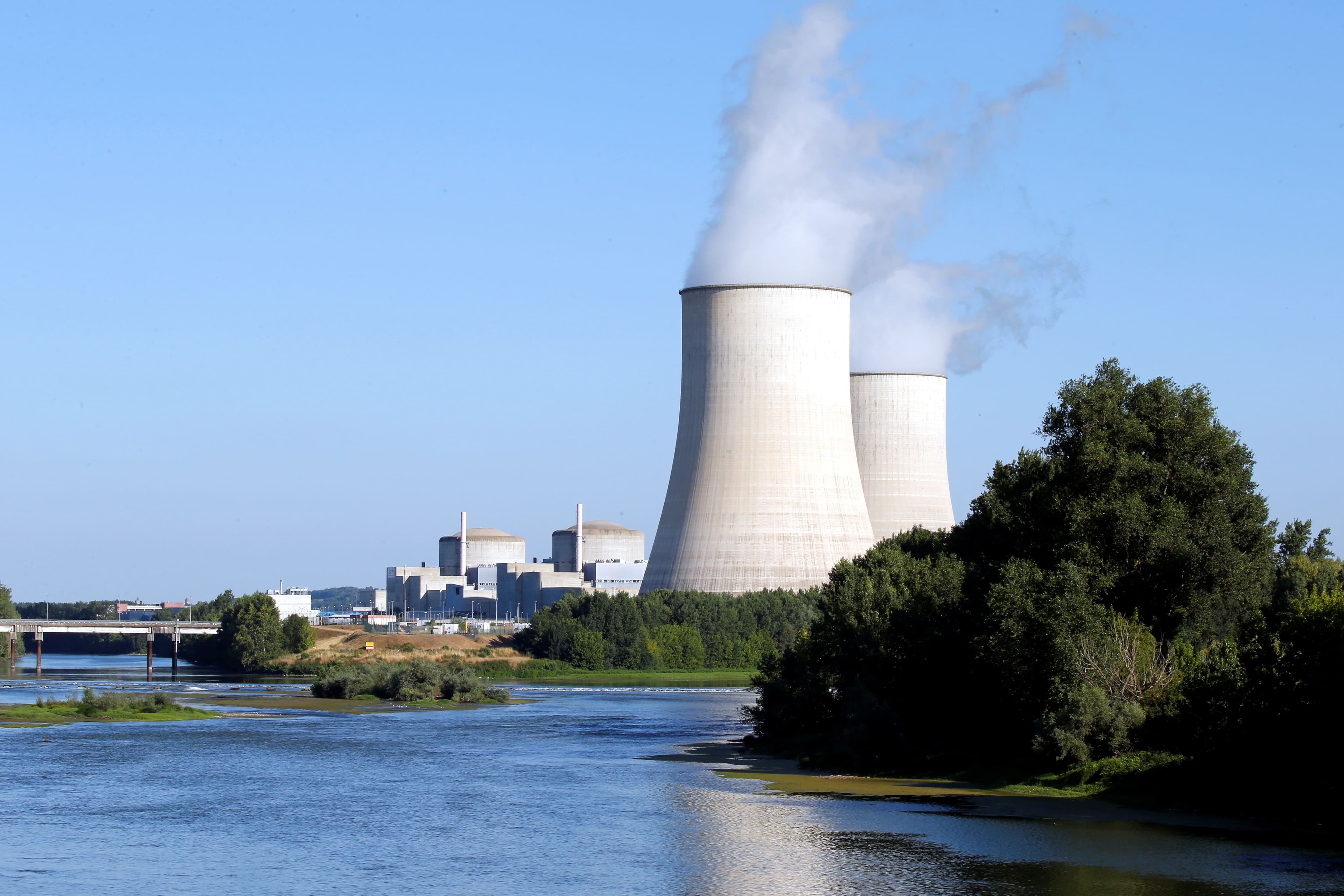 A nuclear power plant with two cooling towers emitting steam, next to a river under a clear sky.