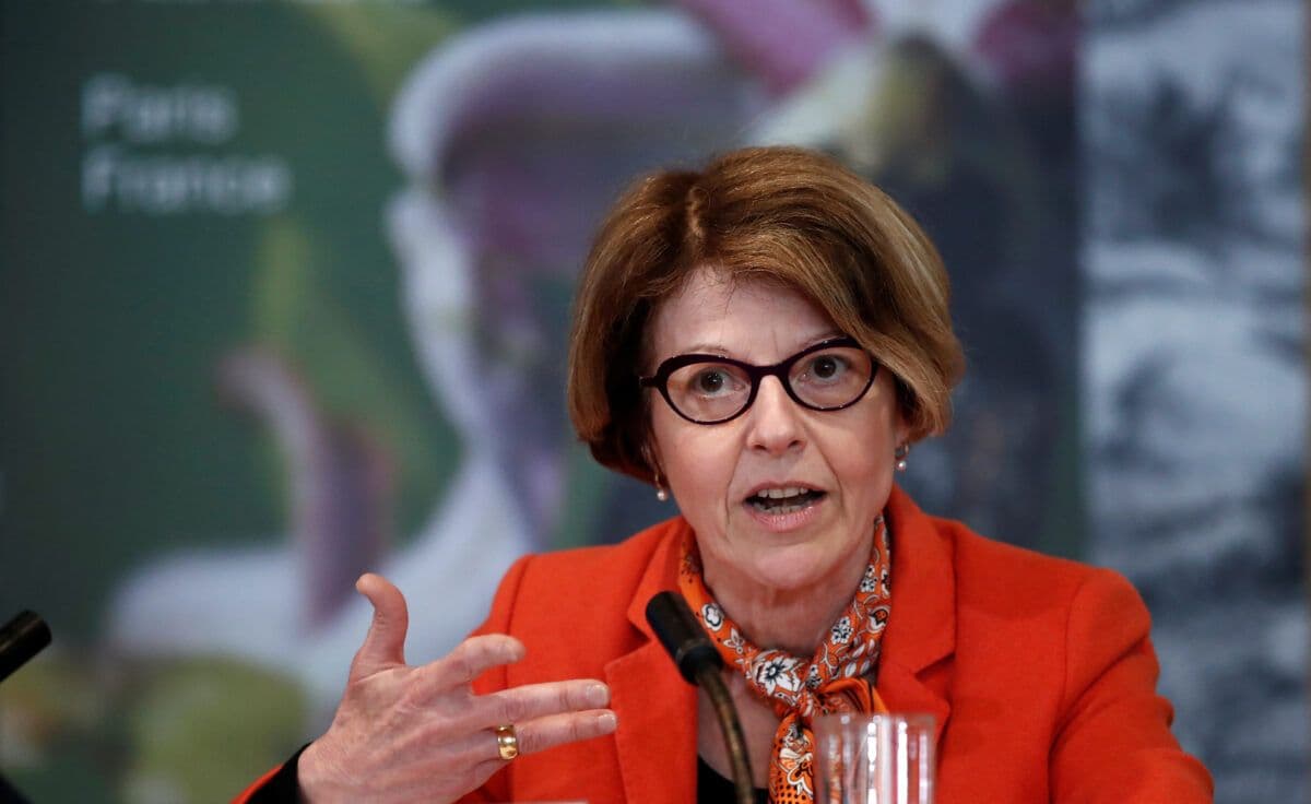 En kvinne med kort brunt hår, iført briller, en rød blazer og et oransje skjerf, snakker animert ved en mikrofon under en konferanse.