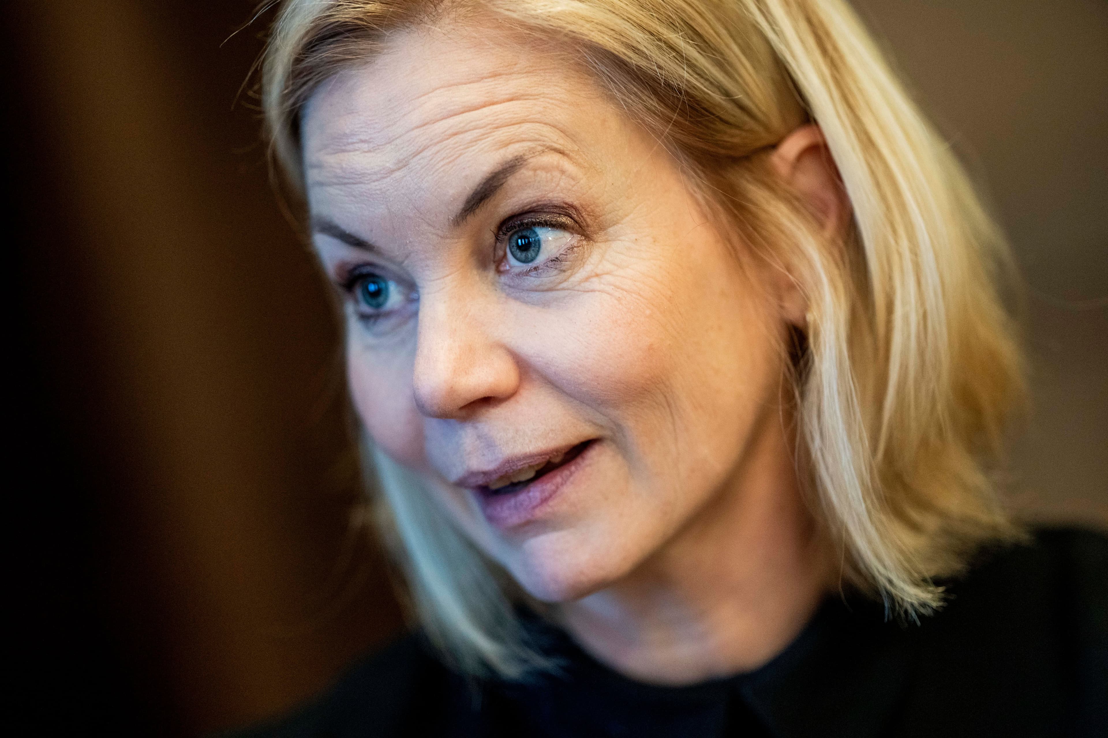 En blond kvinne i en svart kjole går smilende på en rød løper, med en svart bil og sikkerhetsvakter i bakgrunnen.