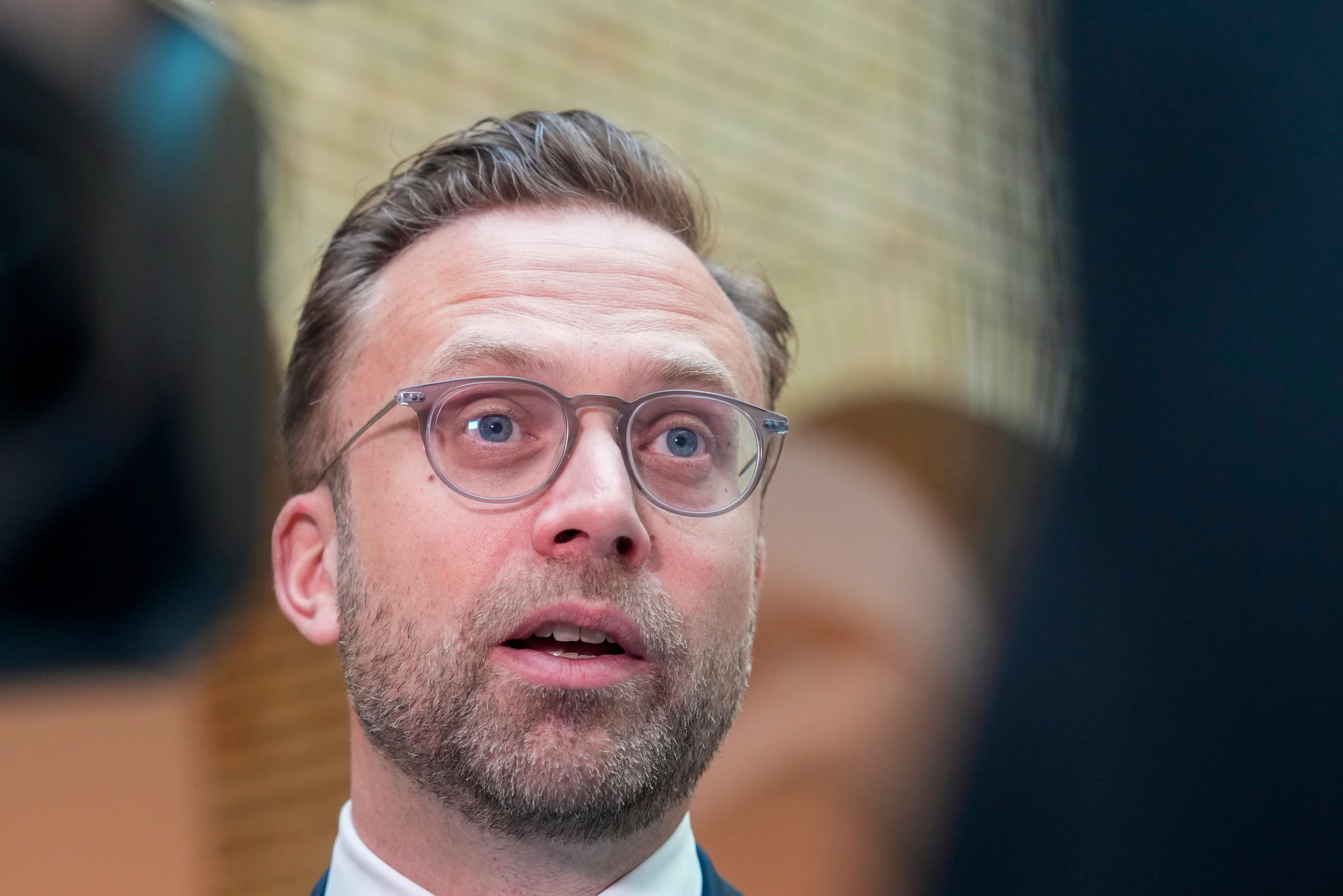 A man with glasses and a beard looks up with his mouth slightly open, wearing a suit.