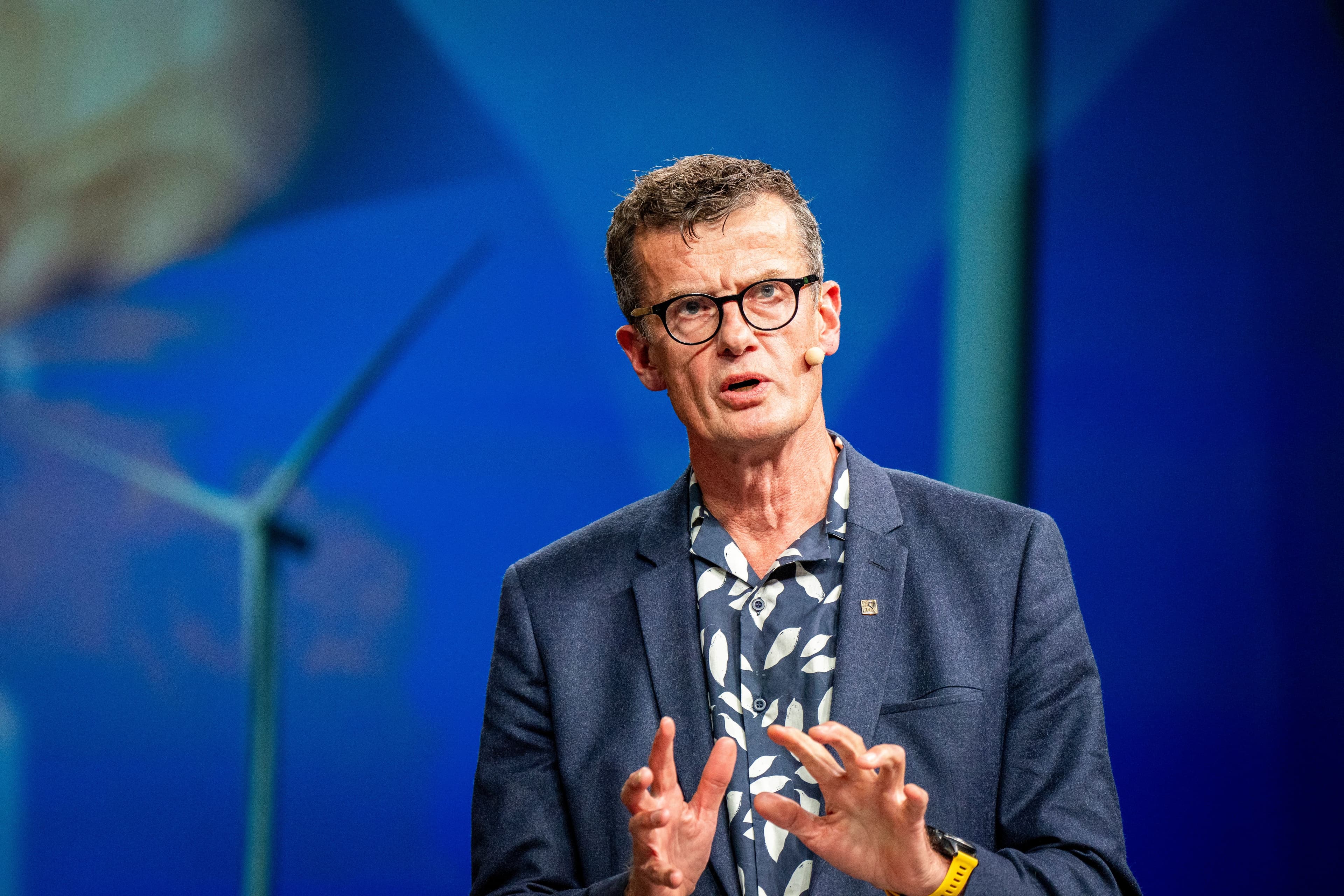 En mann med briller, blazer og mønstret skjorte snakker og gestikulerer med hendene foran en uskarp blå bakgrunn med silhuetten av en vindturbin.