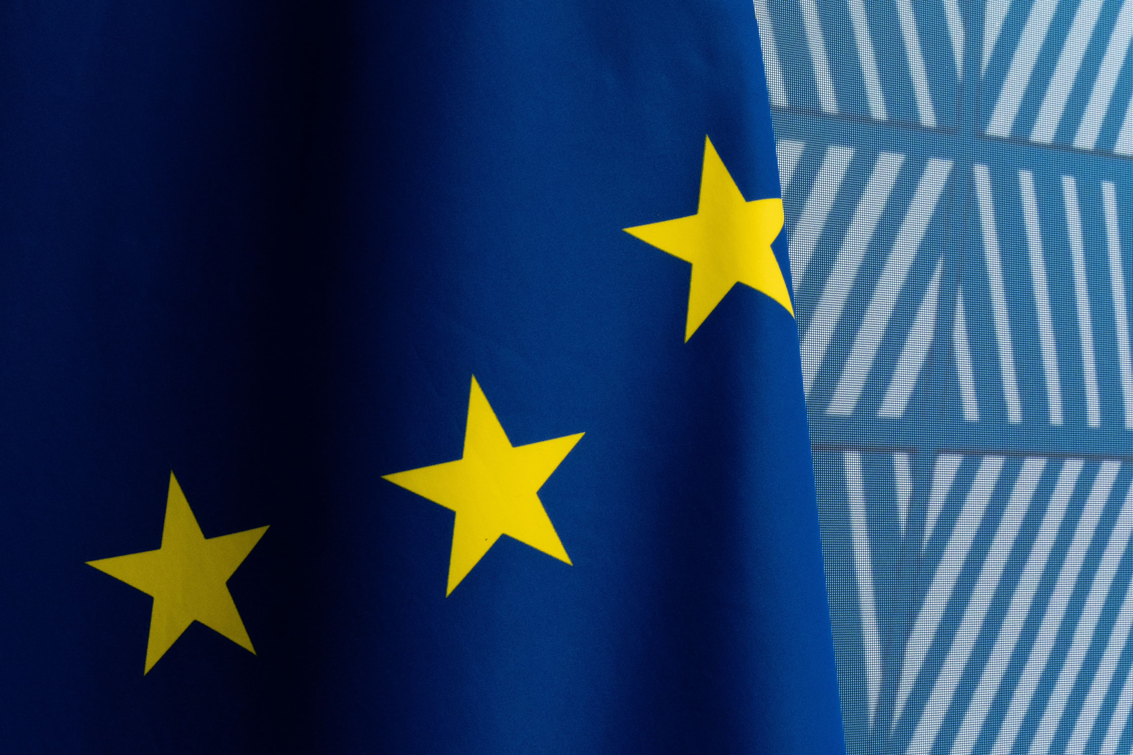 Utsnitt av EU-flagget med tre gylne stjerner på blå bakgrunn, ved siden av en mønstret blå vegg.