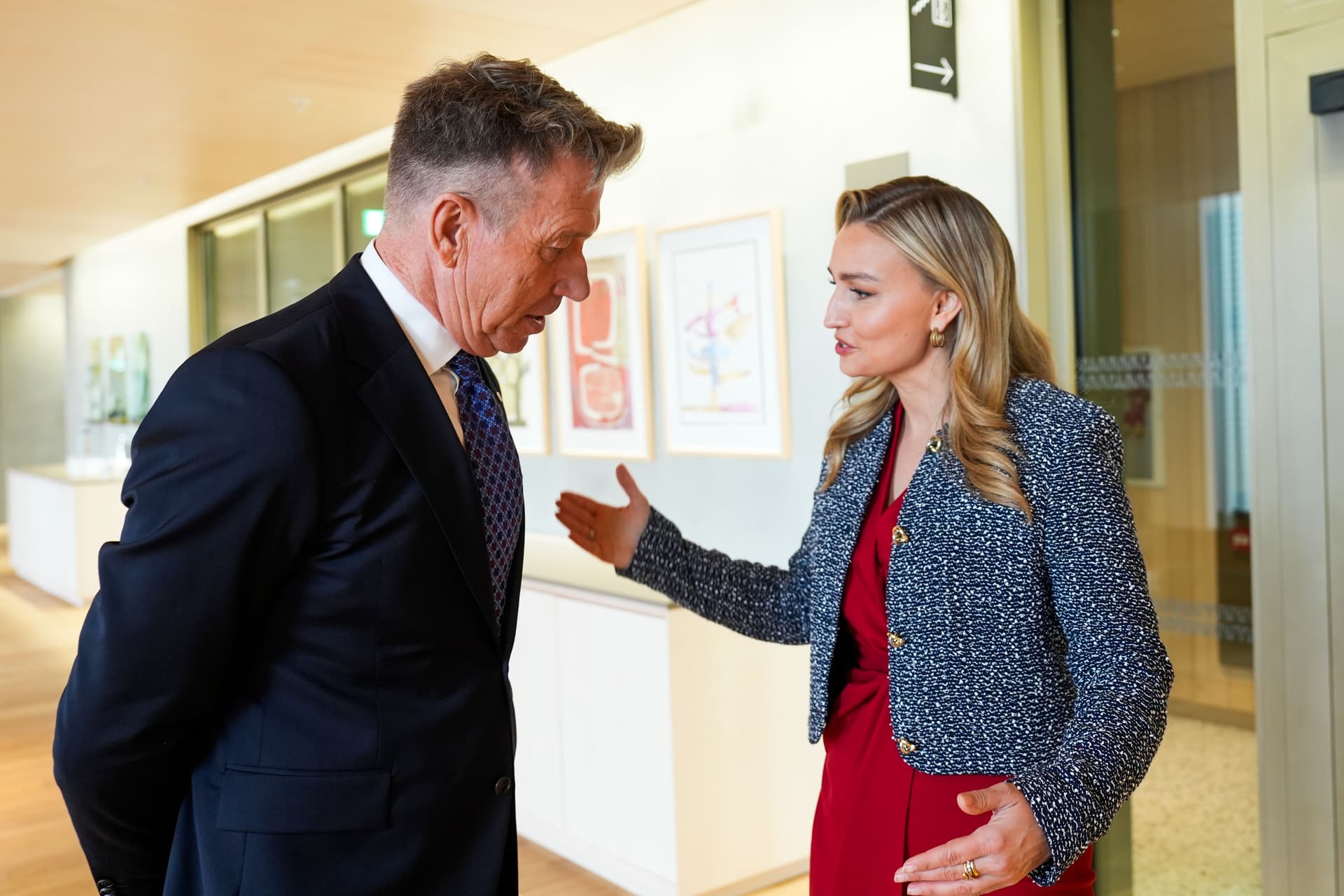 En kvinne i rød kjole og blå jakke gestikulerer mens hun snakker med en mann i mørk dress.
