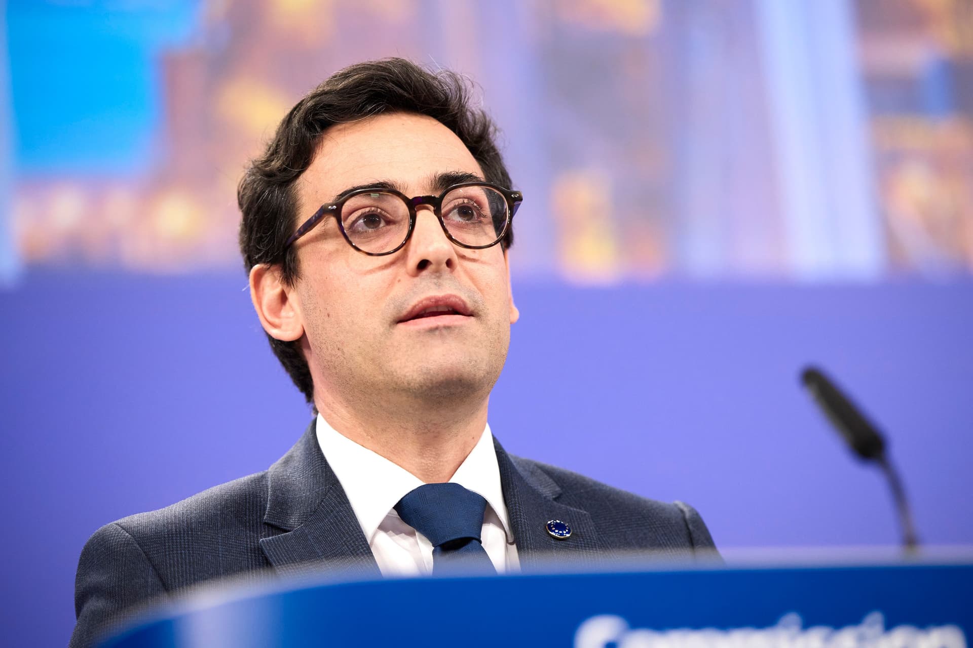 En mann med mørkt hår og briller i dress og slips taler fra en talerstol.