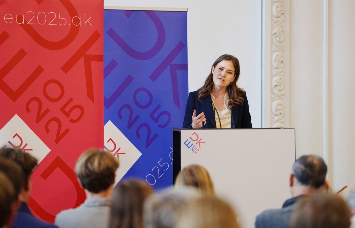 En kvinne står på et podium og taler til et publikum. Bak henne henger røde og blå bannere med "DK 2025" og nettstedet "eu2025.dk".