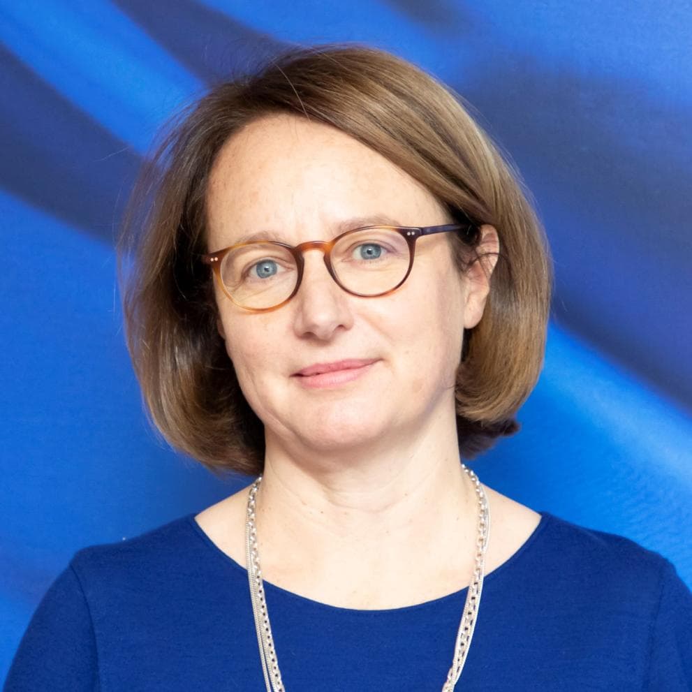 Portrettbilde av en kvinne med kort brunt hår og briller, iført en blå topp, mot en blå bakgrunn.